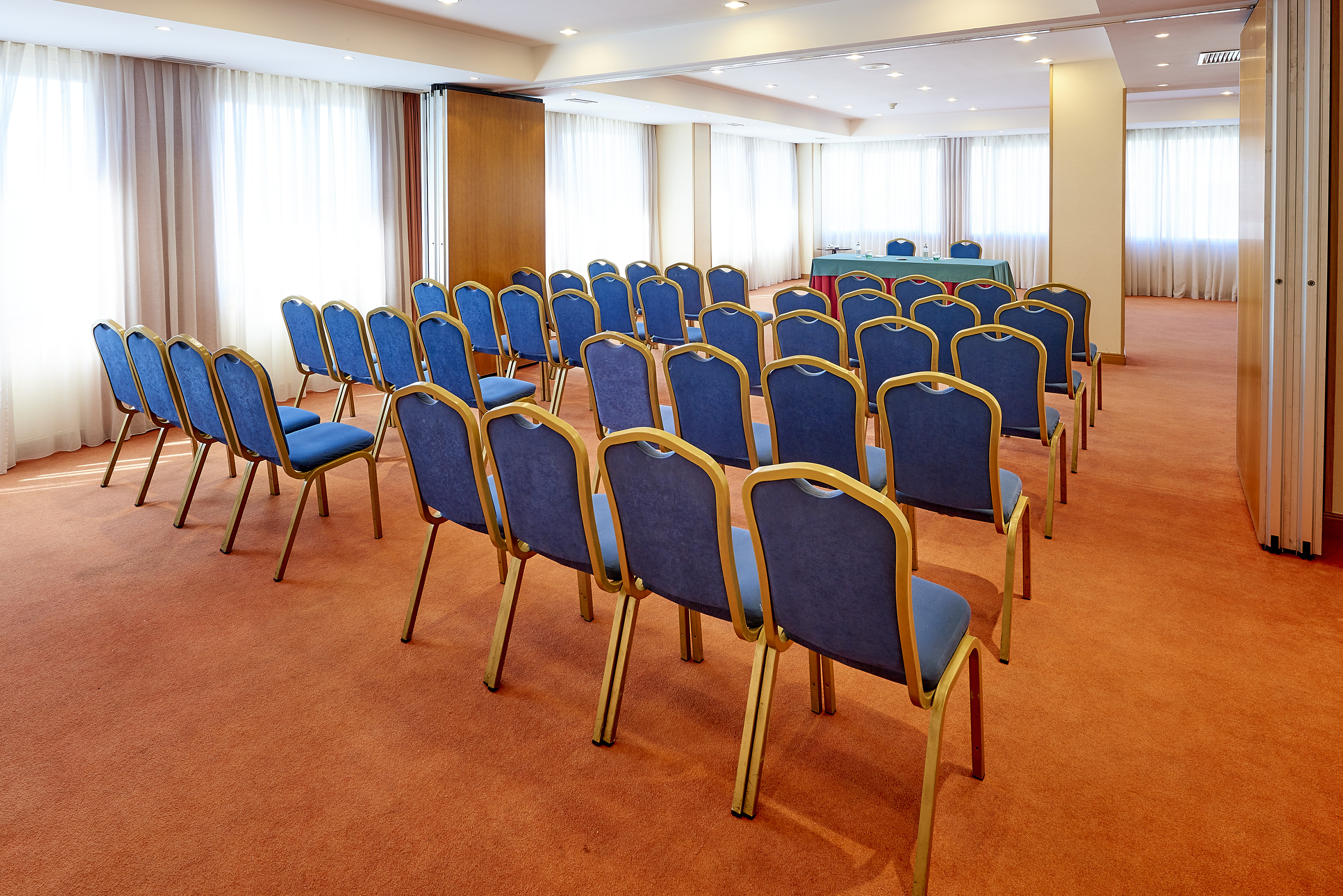 Empty conference room with rows of blue chairs, orange carpet, and white curtains.