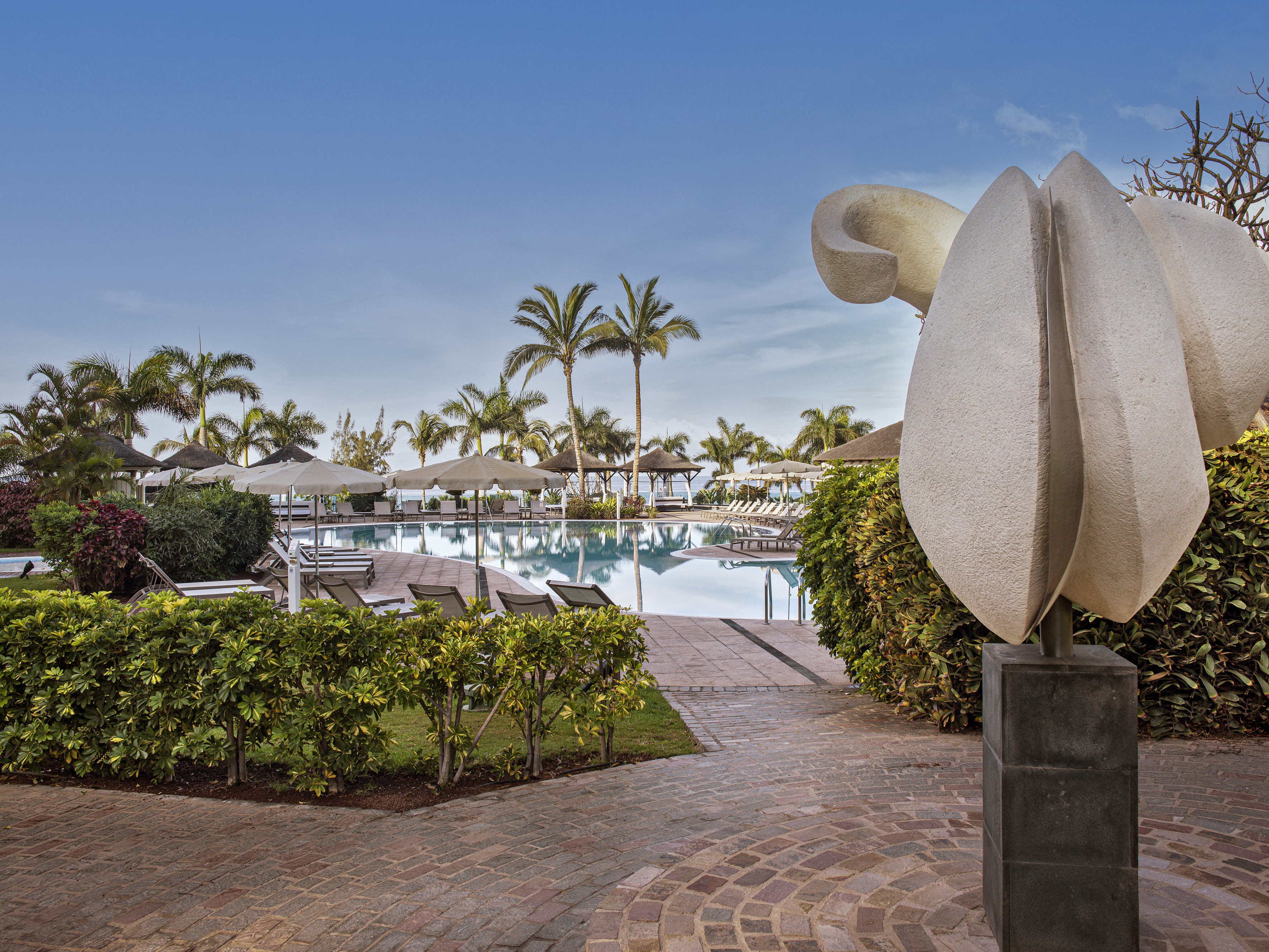 Tropical resort swimming pool with palm trees, modern sculpture, and brick pathway.