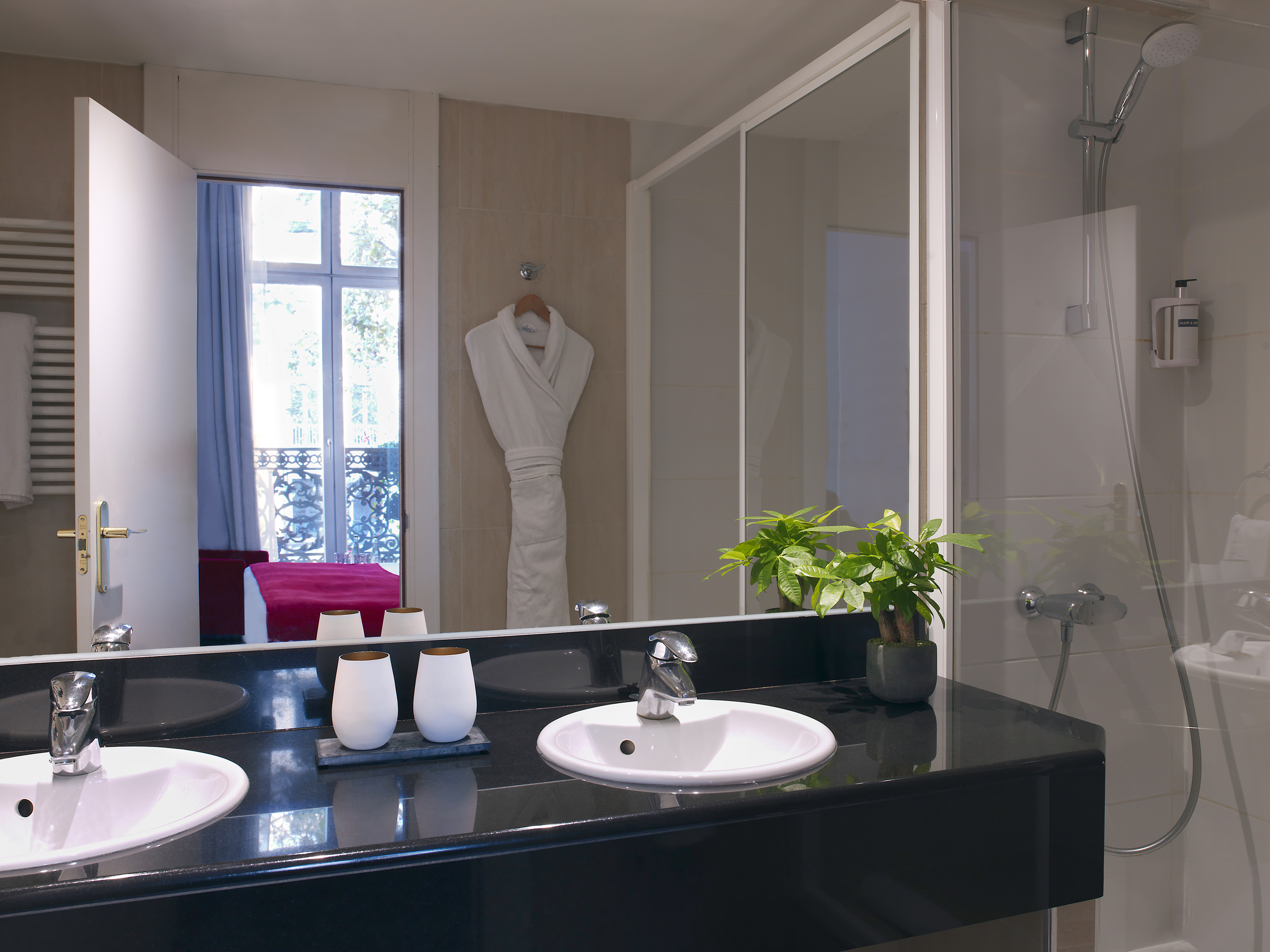 Modern hotel bathroom with two sinks, glass shower, and mirror reflecting a bedroom.