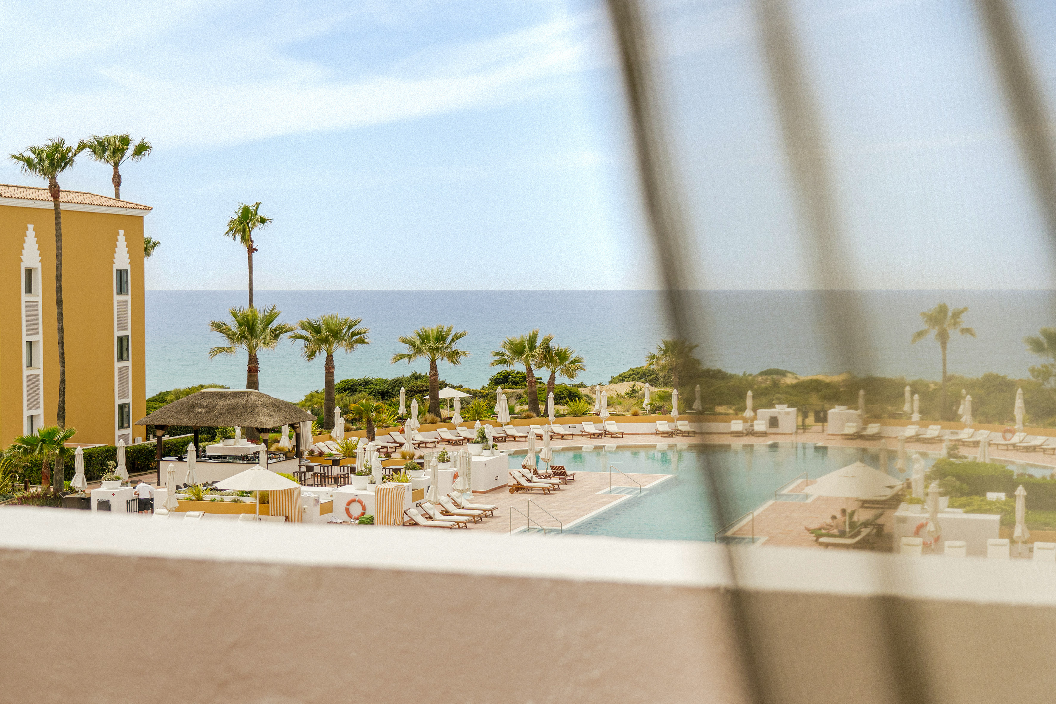 Tropical resort pool area with ocean view, palm trees, and yellow hotel building.