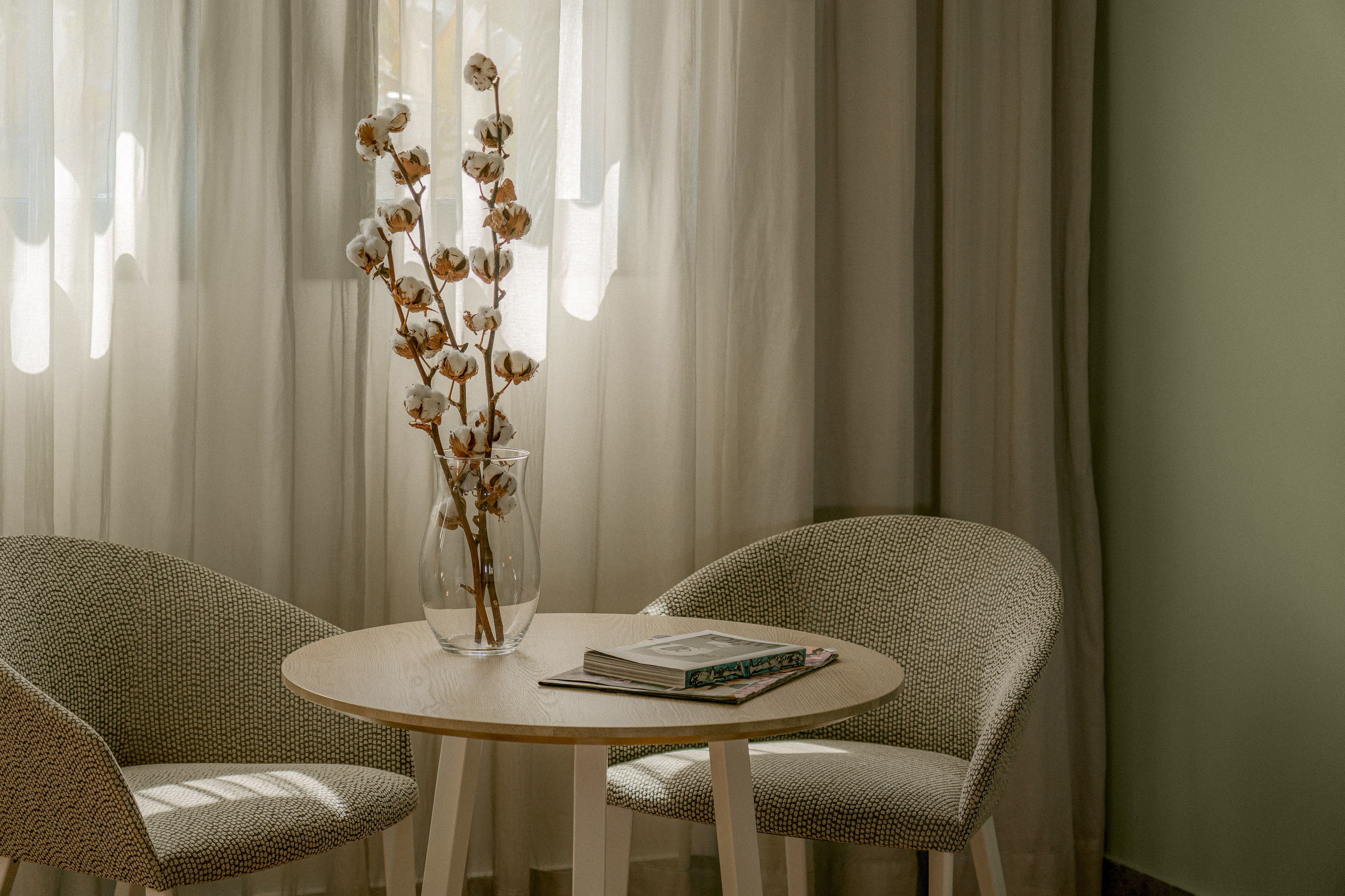 Cotton branches in vase, round wooden table, two chairs, and magazine.