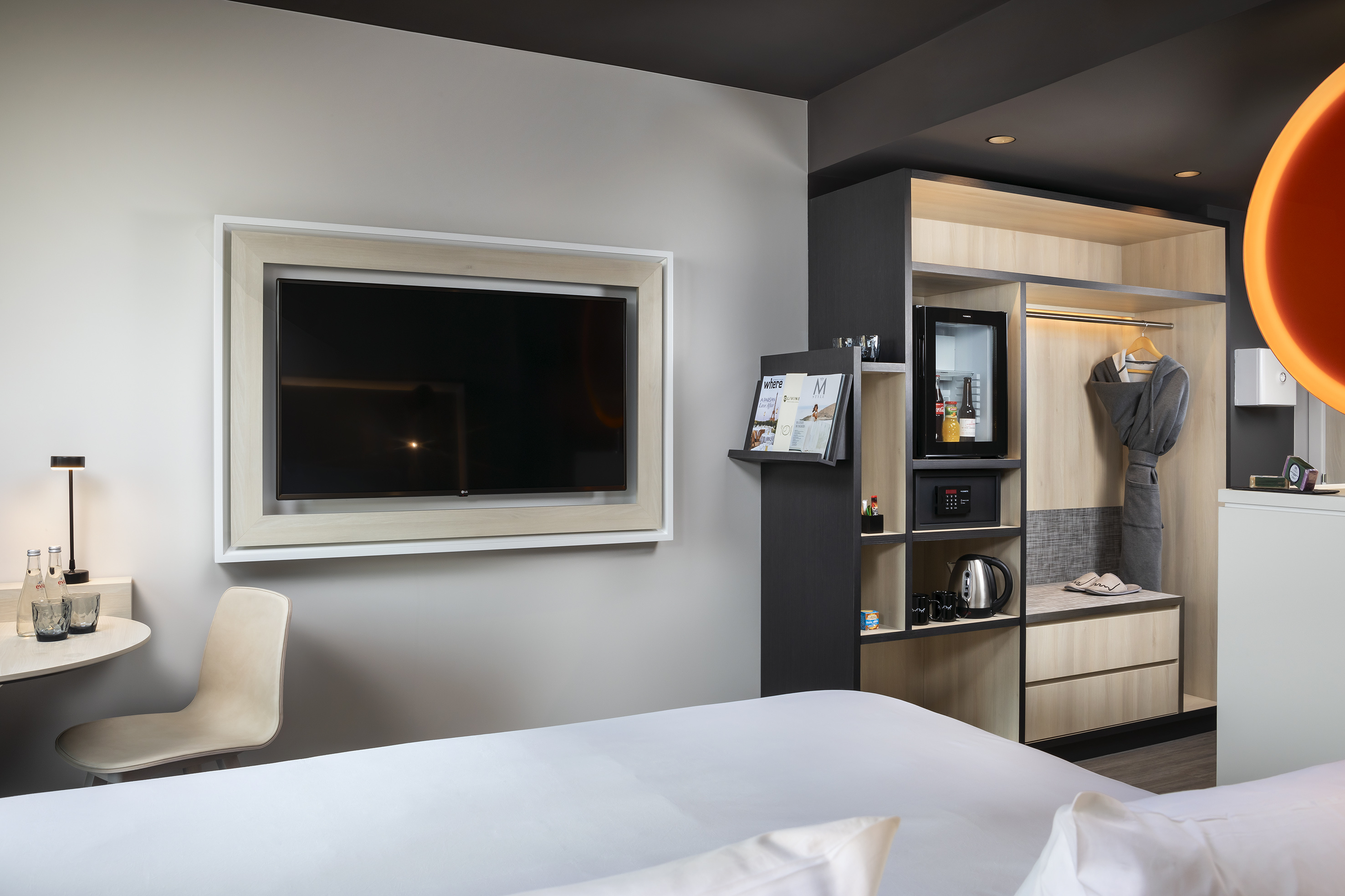 Modern hotel room with wall-mounted TV, wardrobe, minibar, and desk area.