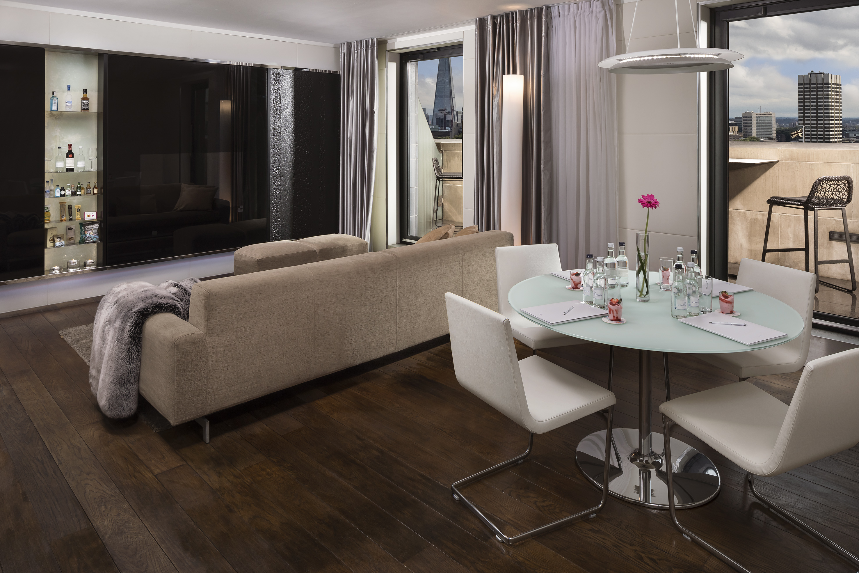 Modern hotel suite with dining table, sofa, built-in bar, and London city view.