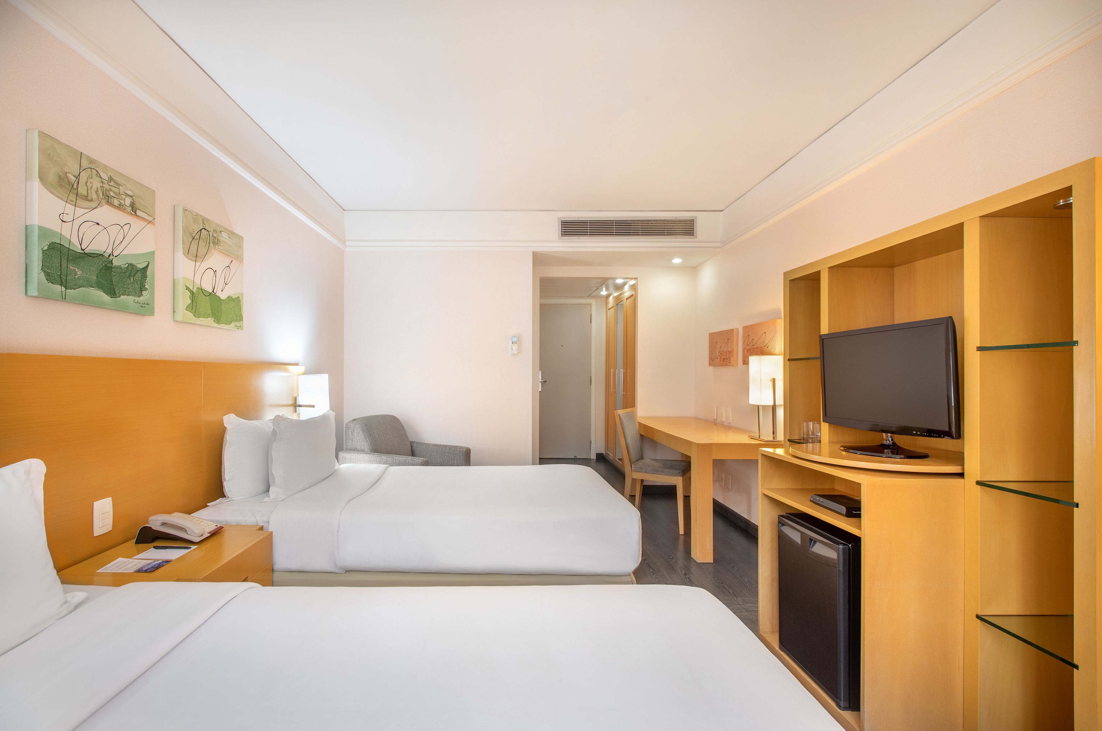 Modern hotel room with two twin beds, desk, flat-screen TV, and mini-fridge.