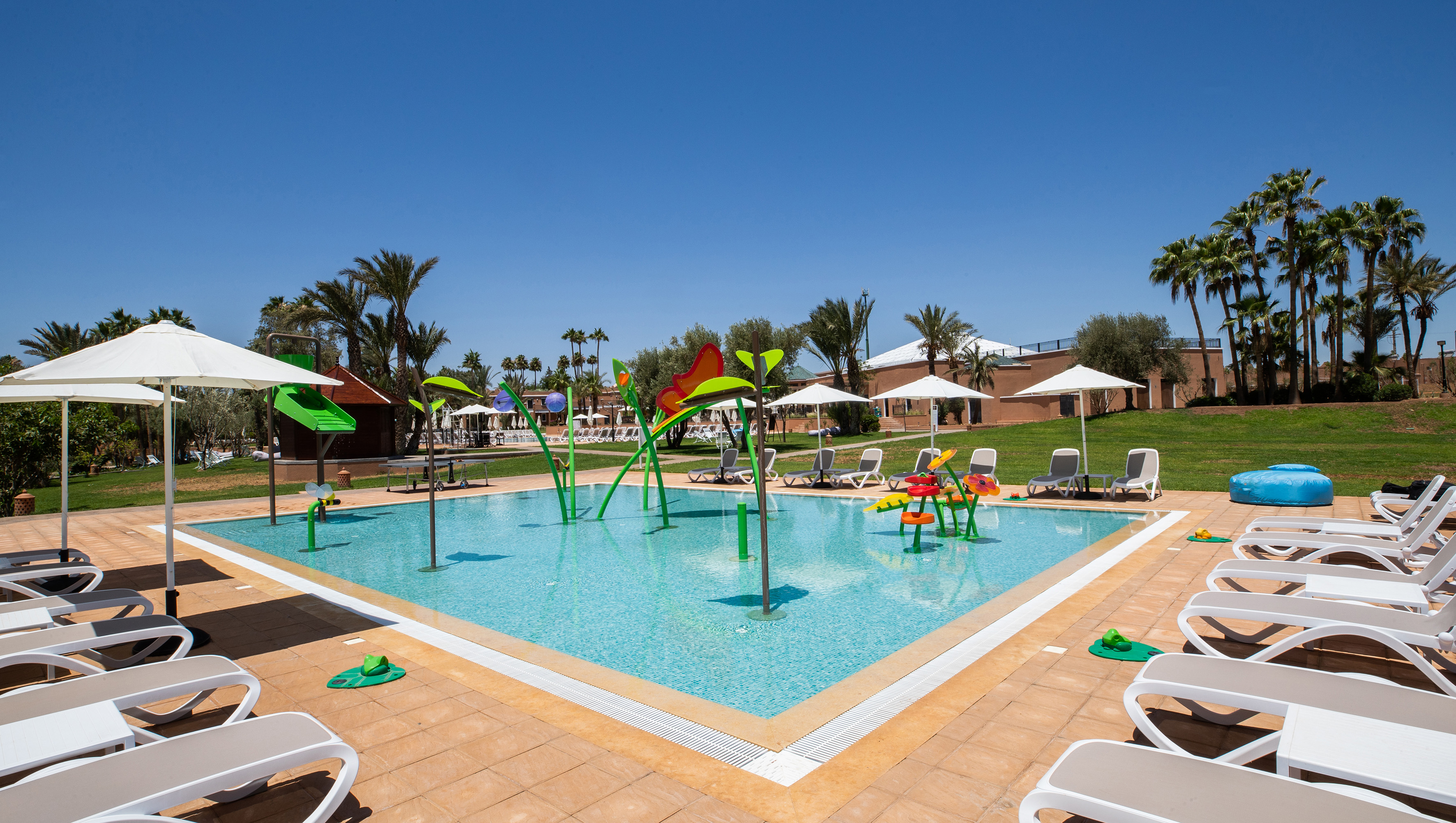 Sunny resort kids splash pad pool with palm trees, lounge chairs, and umbrellas.