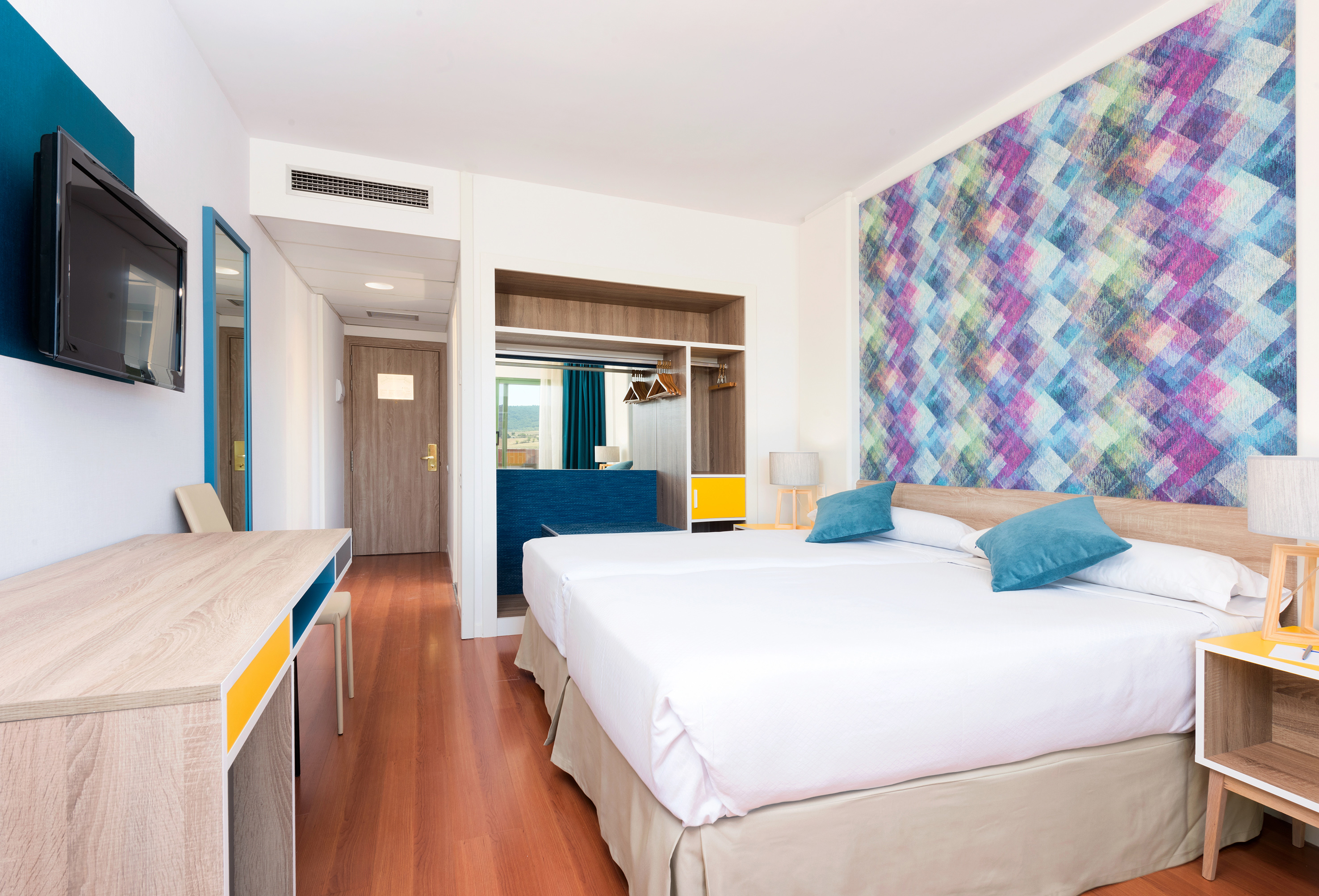 Modern hotel room with twin beds, colorful accent wall, and wood floor.