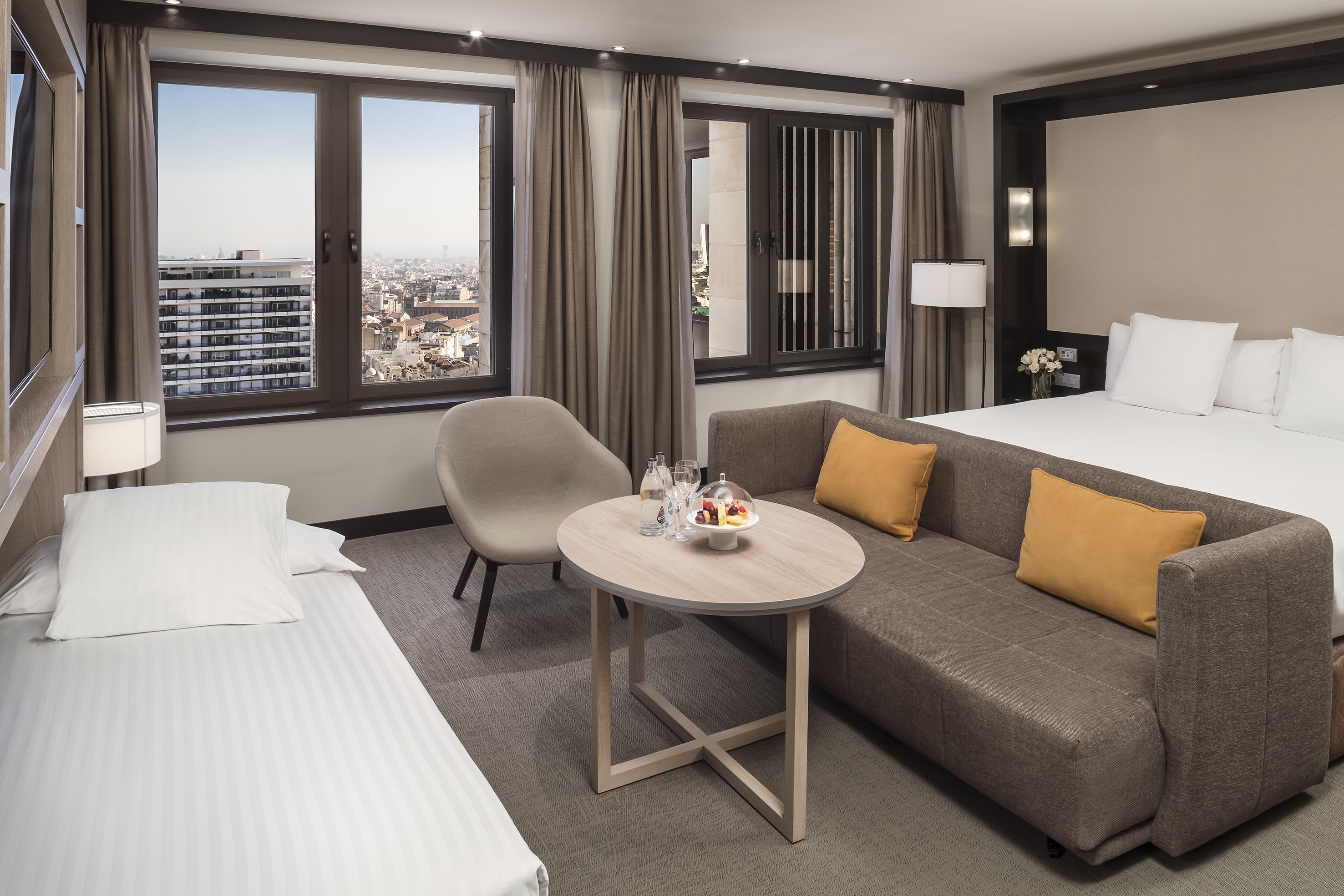 Bright hotel room with city view, large bed, sofa, and single bed.