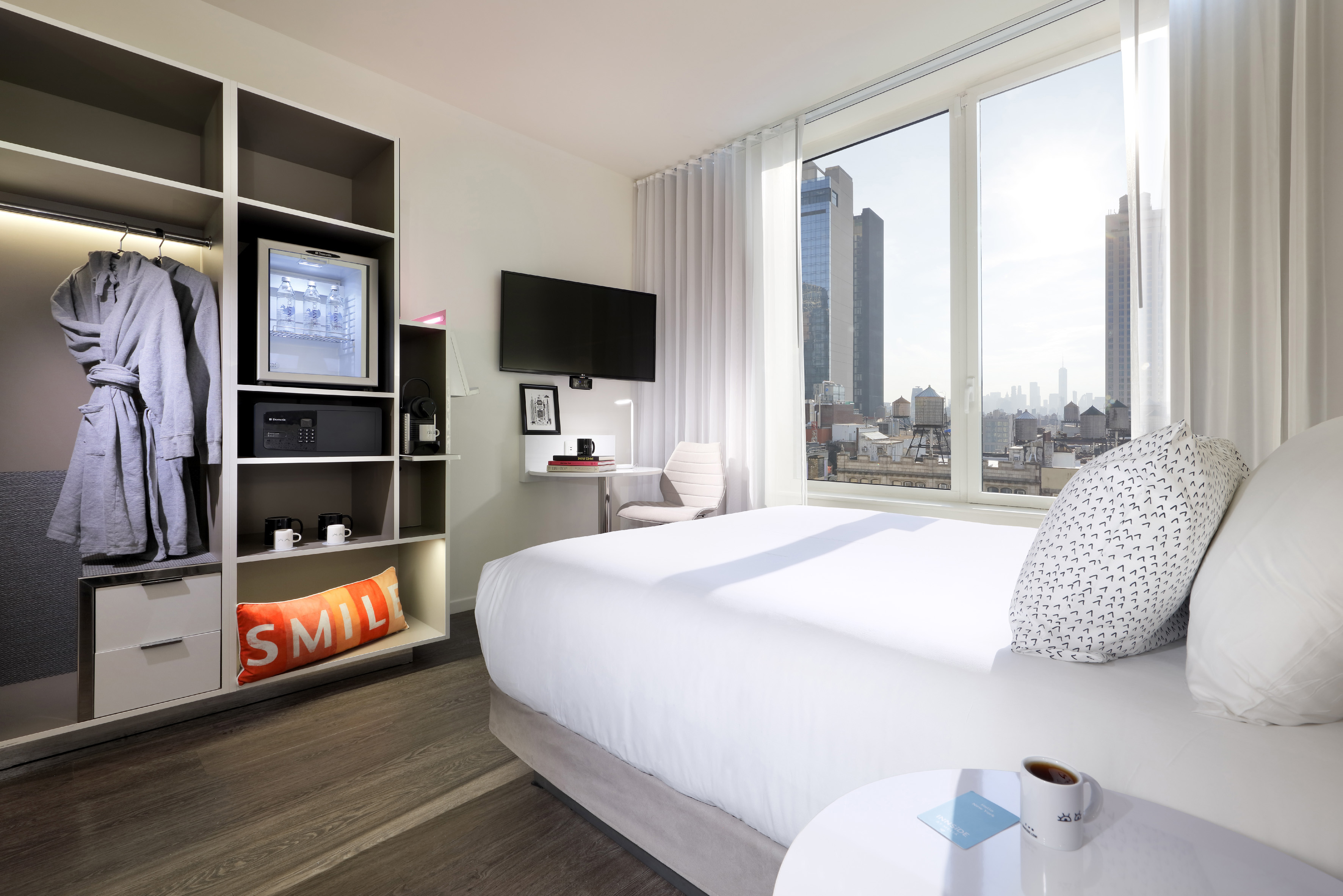 Modern hotel room with New York City view, bed, wardrobe, and mini-fridge.