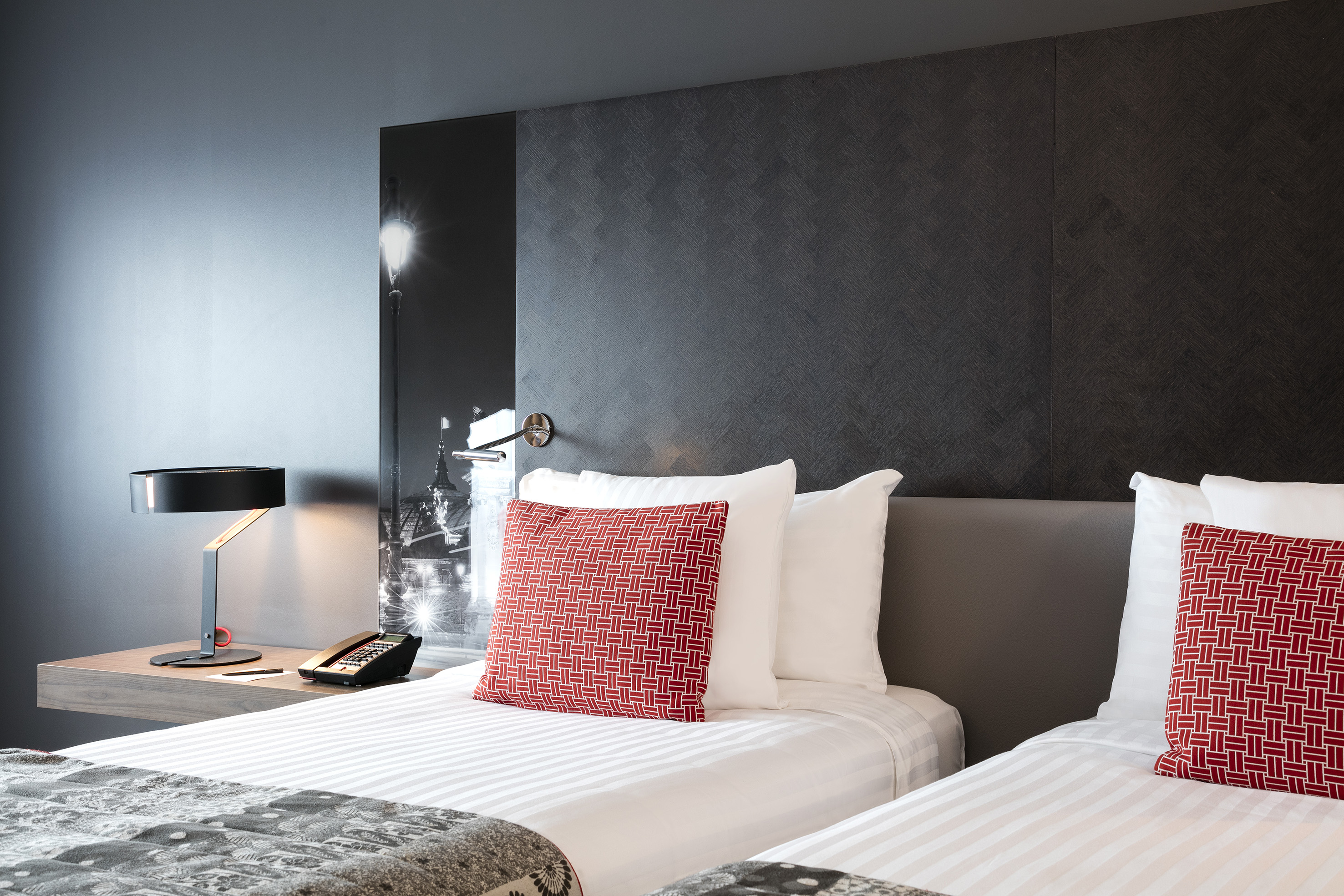 Modern hotel twin room with red pillows, dark wall, and bedside lamp.