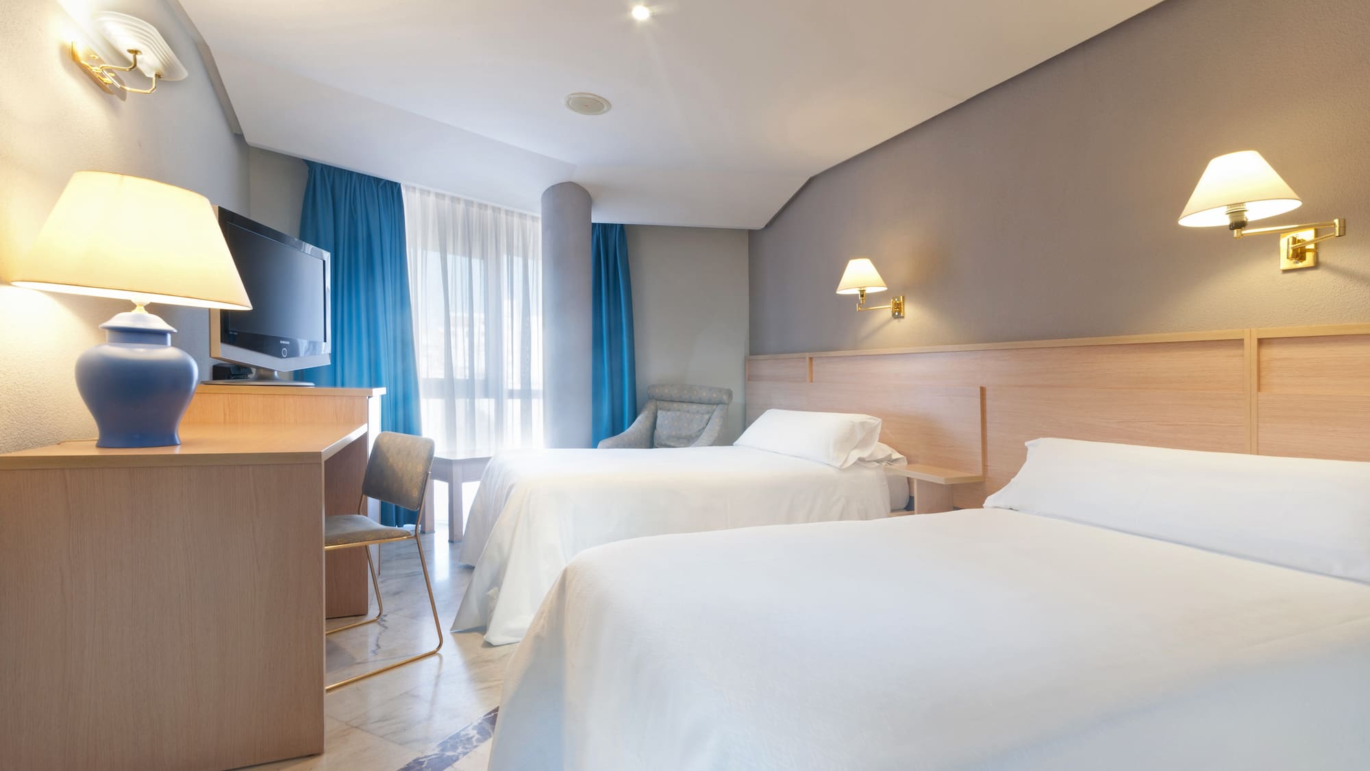 Hotel guest room with two twin beds, work desk, television, and blue curtains.