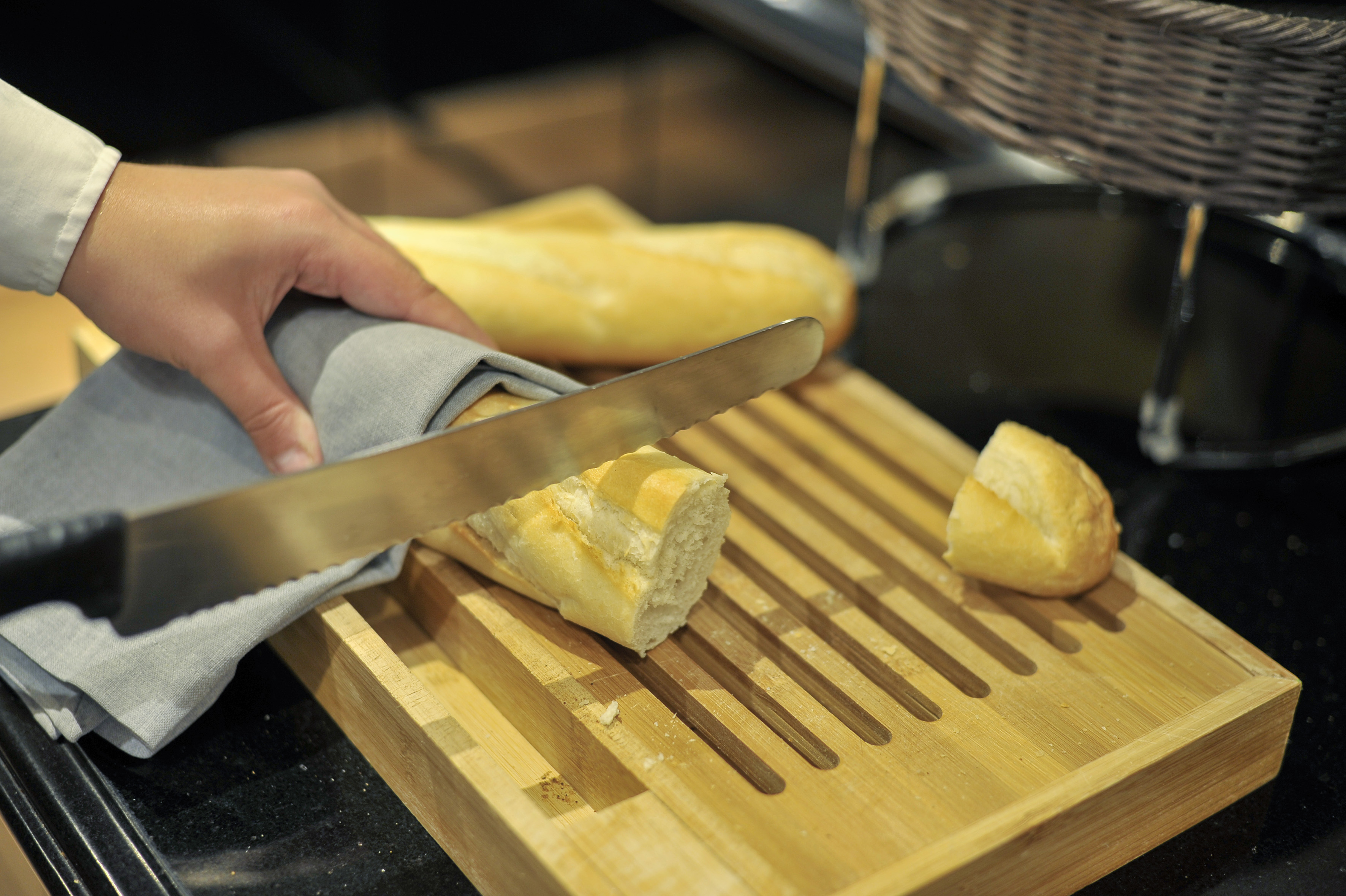 Hand slicing fresh baguette on a wooden cutting board with a bread knife.