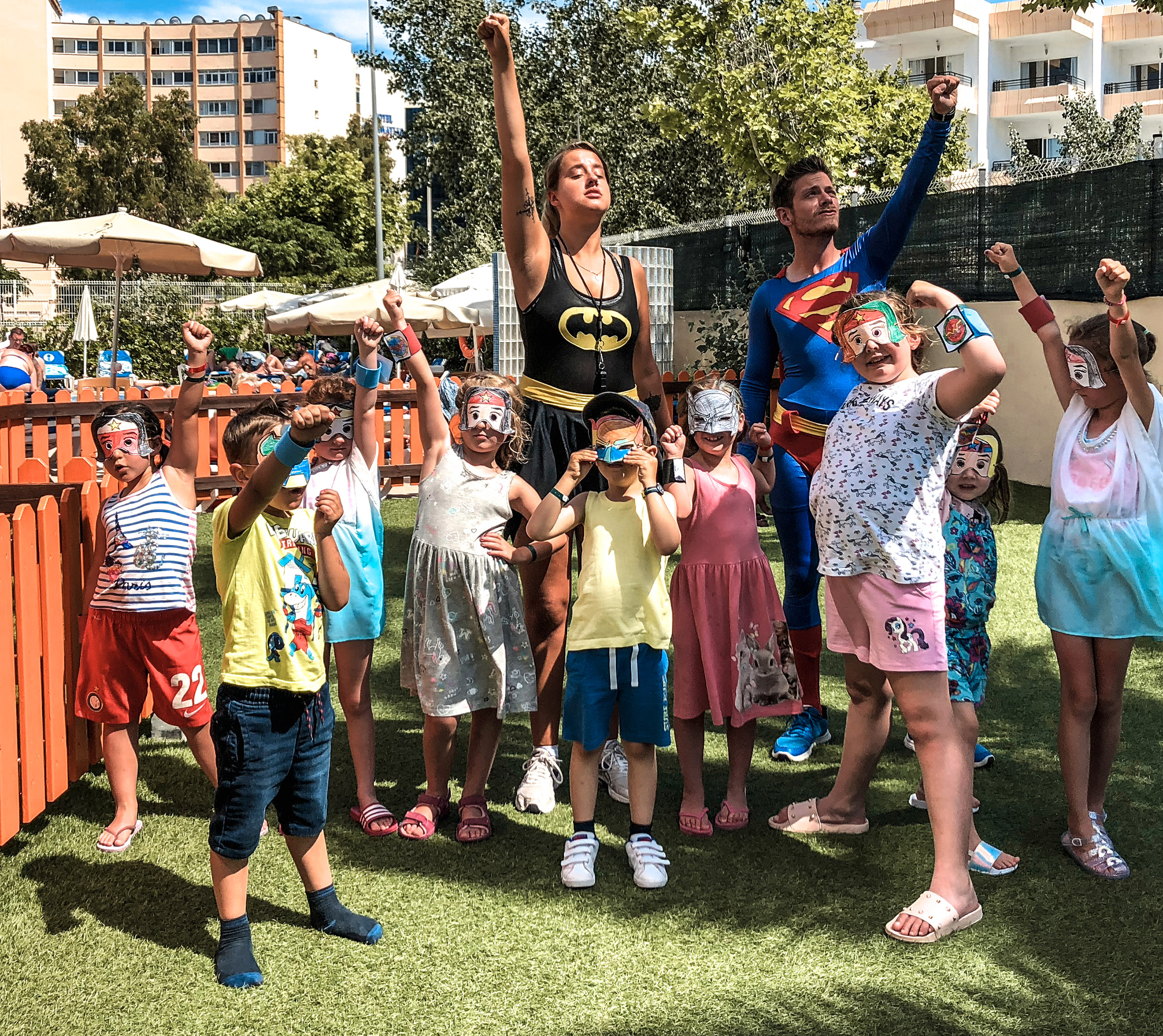 Kids and costumed adults (Batman, Superman) with raised fists at superhero party on grass.