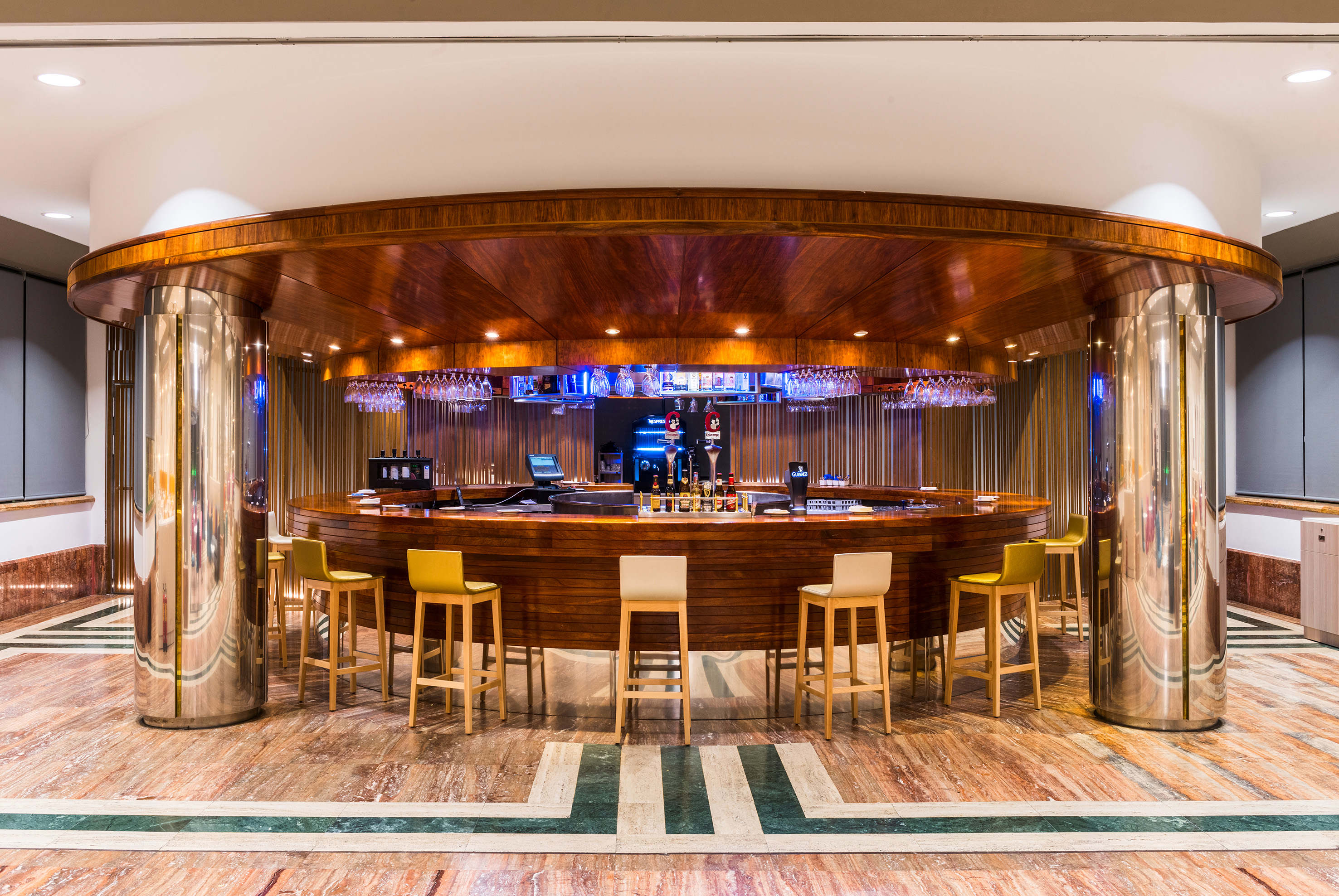 Circular wooden hotel bar with yellow stools and marble floor.