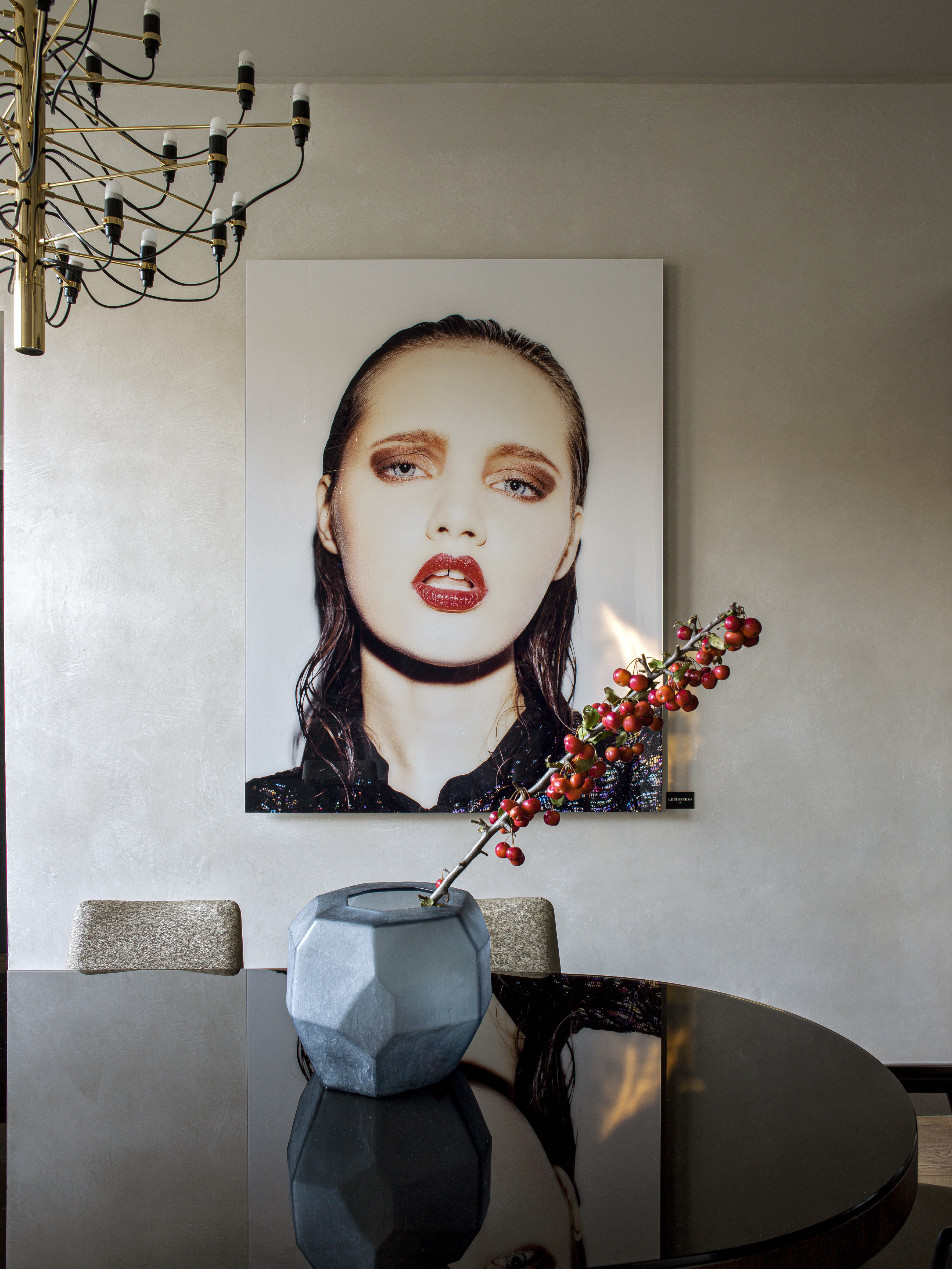 Dining room with modern portrait art, brass chandelier, and berries in grey vase.