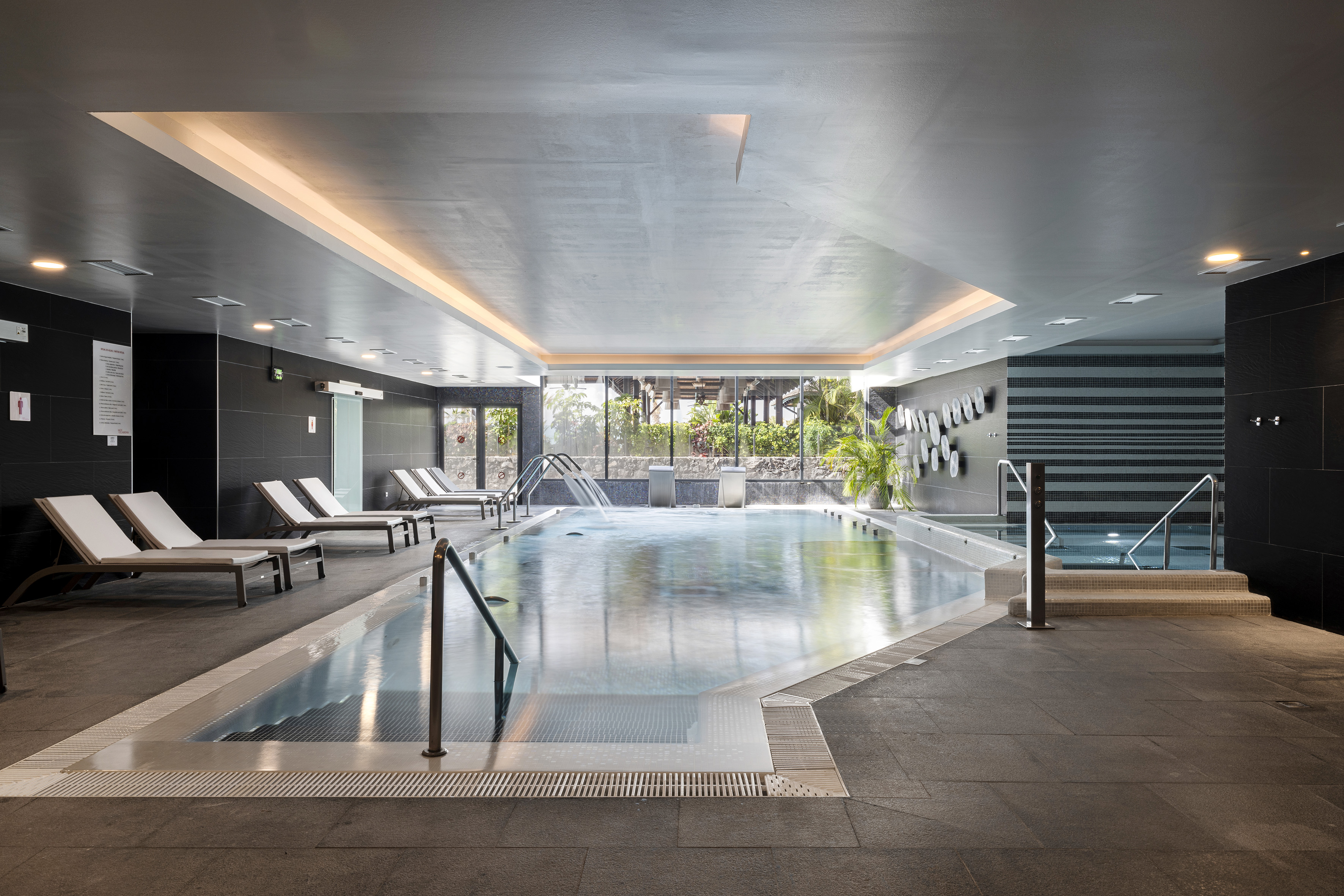 Modern indoor pool with lounge chairs, waterfalls, and garden window views.