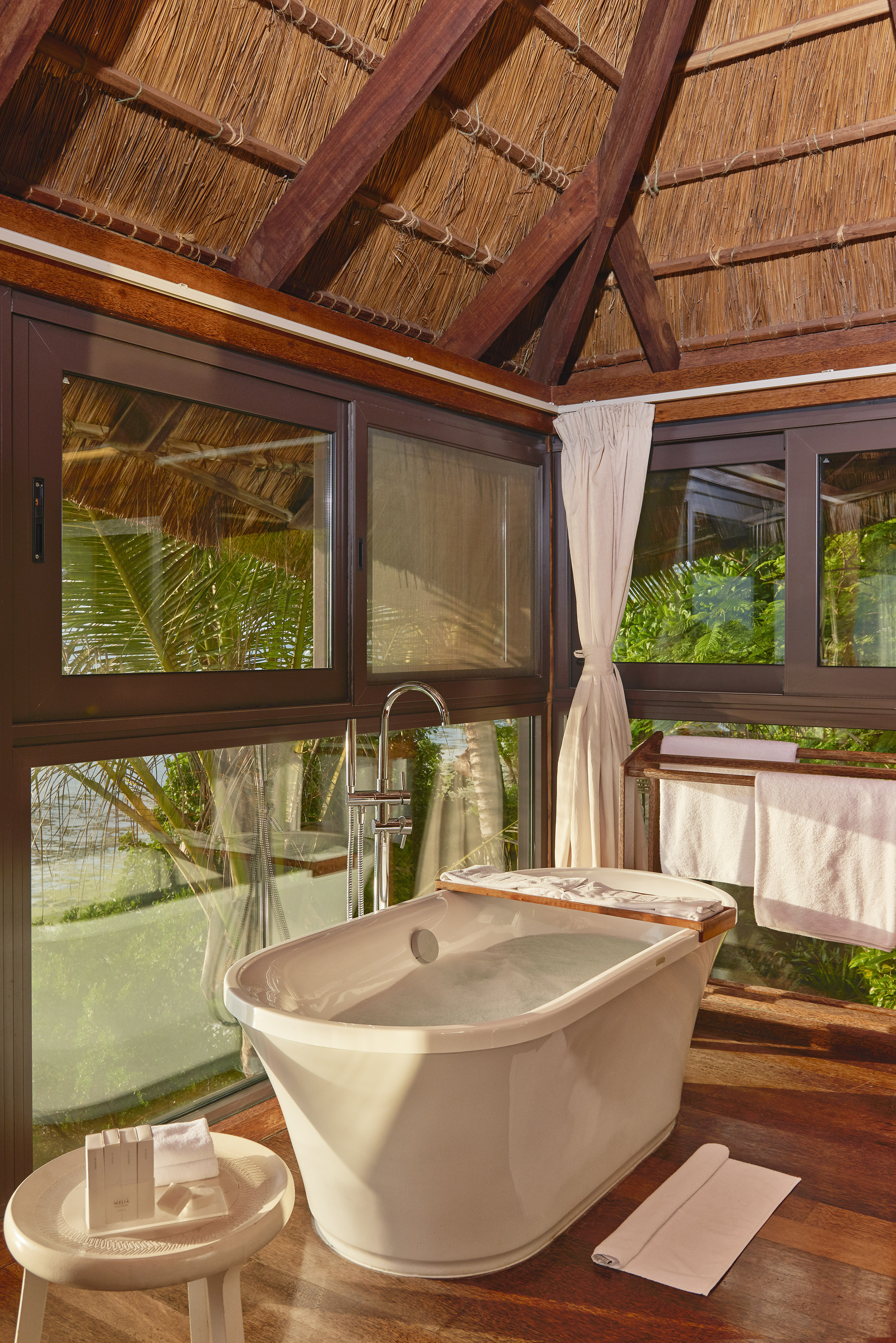 Luxury tropical bathroom with freestanding tub, thatched roof, and scenic view.