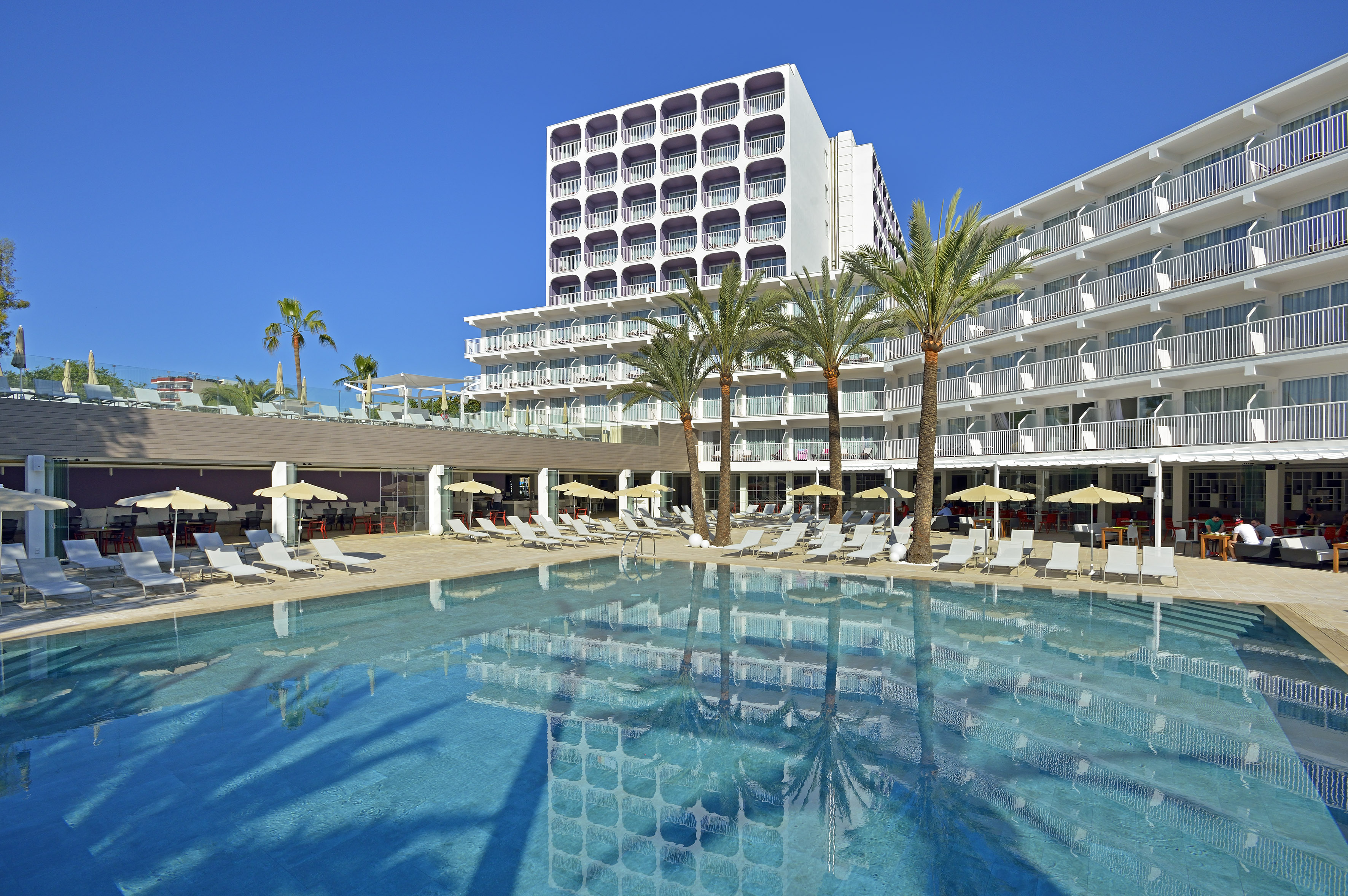 Modern resort hotel with large pool, palm trees, and bright blue sky.