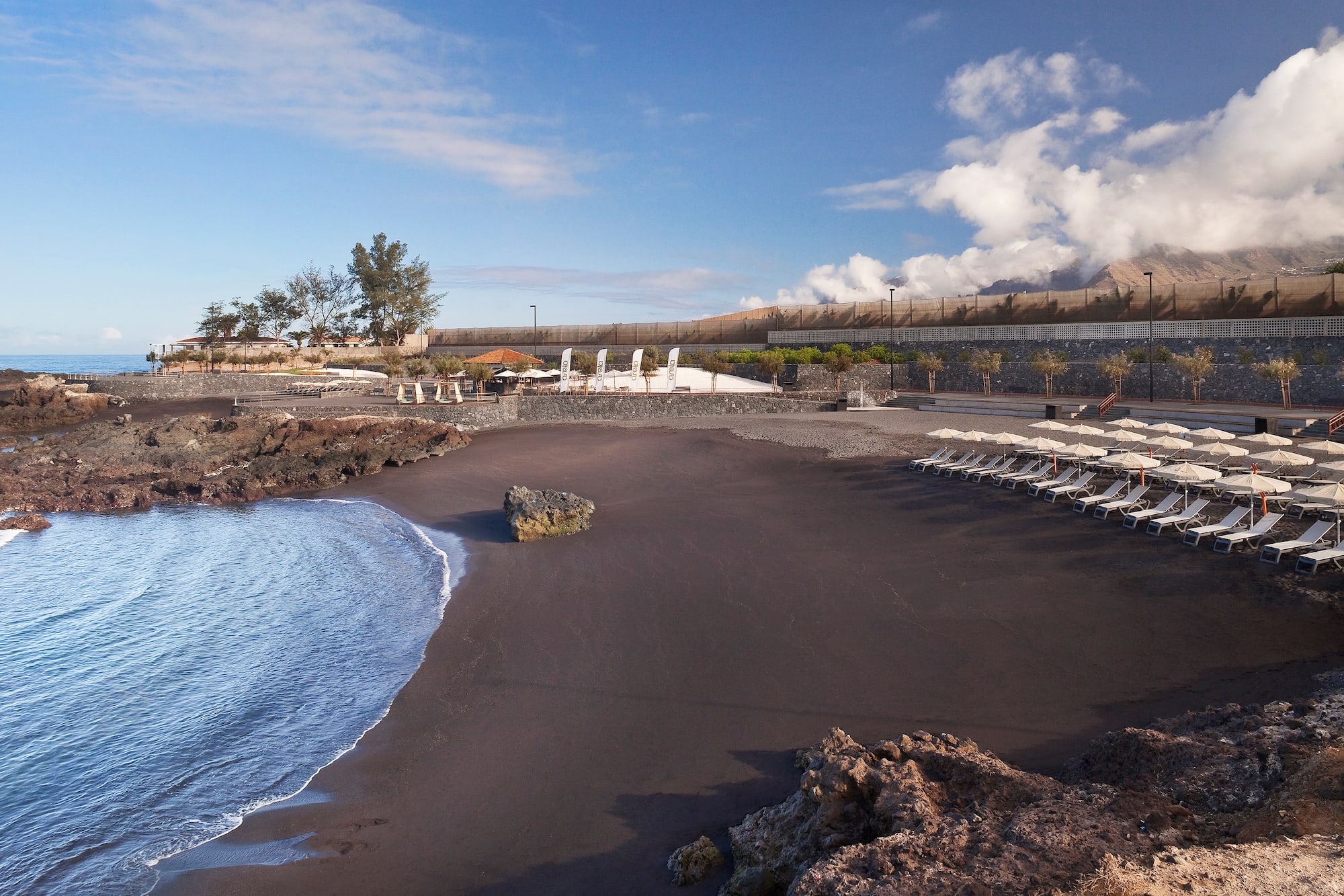 Volcanic black sand beach, lounge chairs, ocean, rocks, and cloudy mountains.