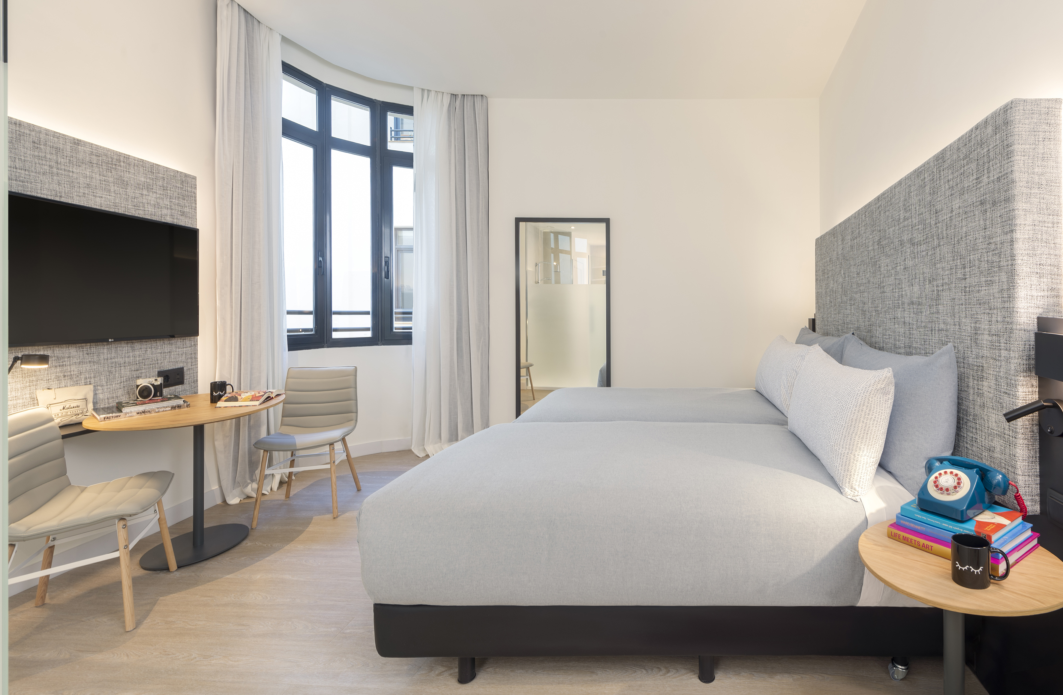 Modern hotel room with grey twin beds, desk, TV, and large window.