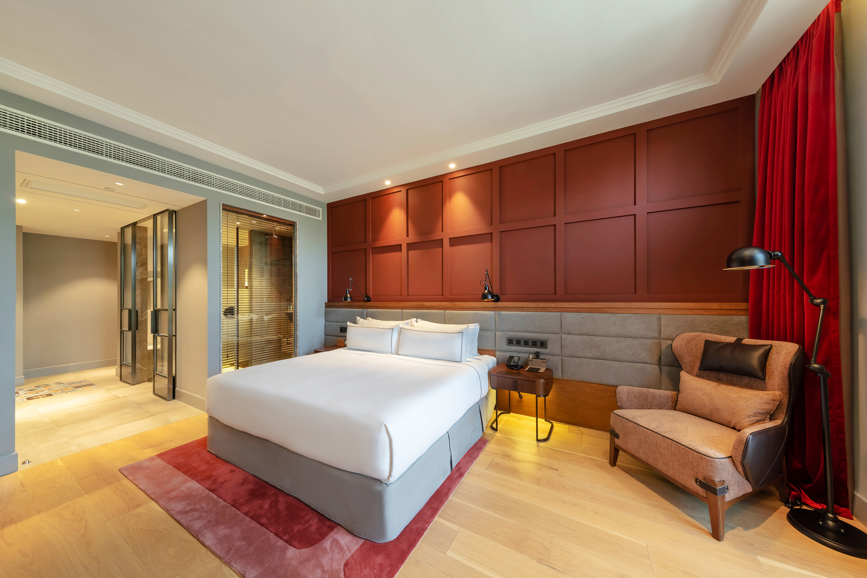 Luxury hotel bedroom with king bed, red wall paneling, and cozy armchair.