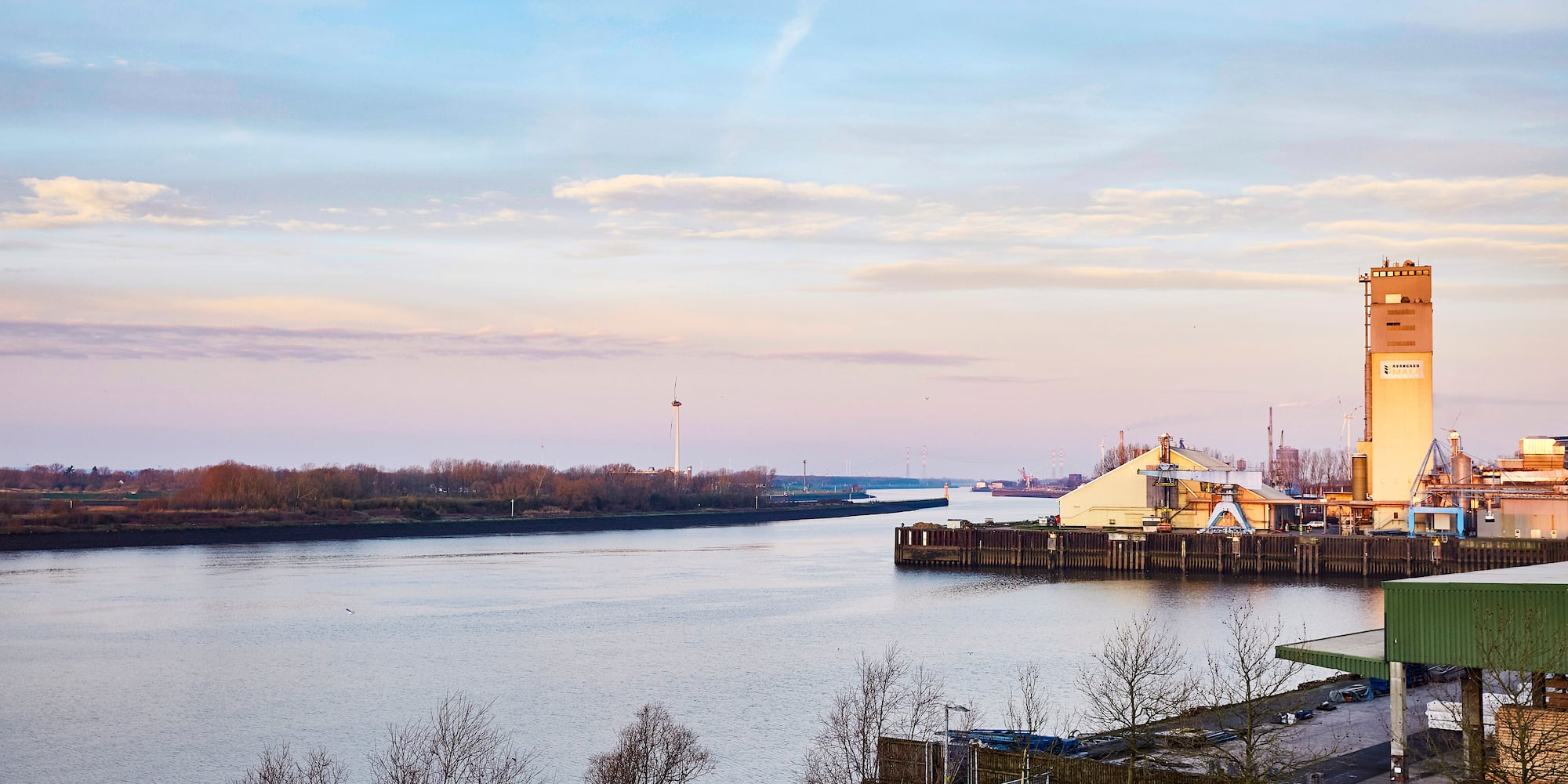 River port with industrial facilities, silo, and bare trees under soft sky.