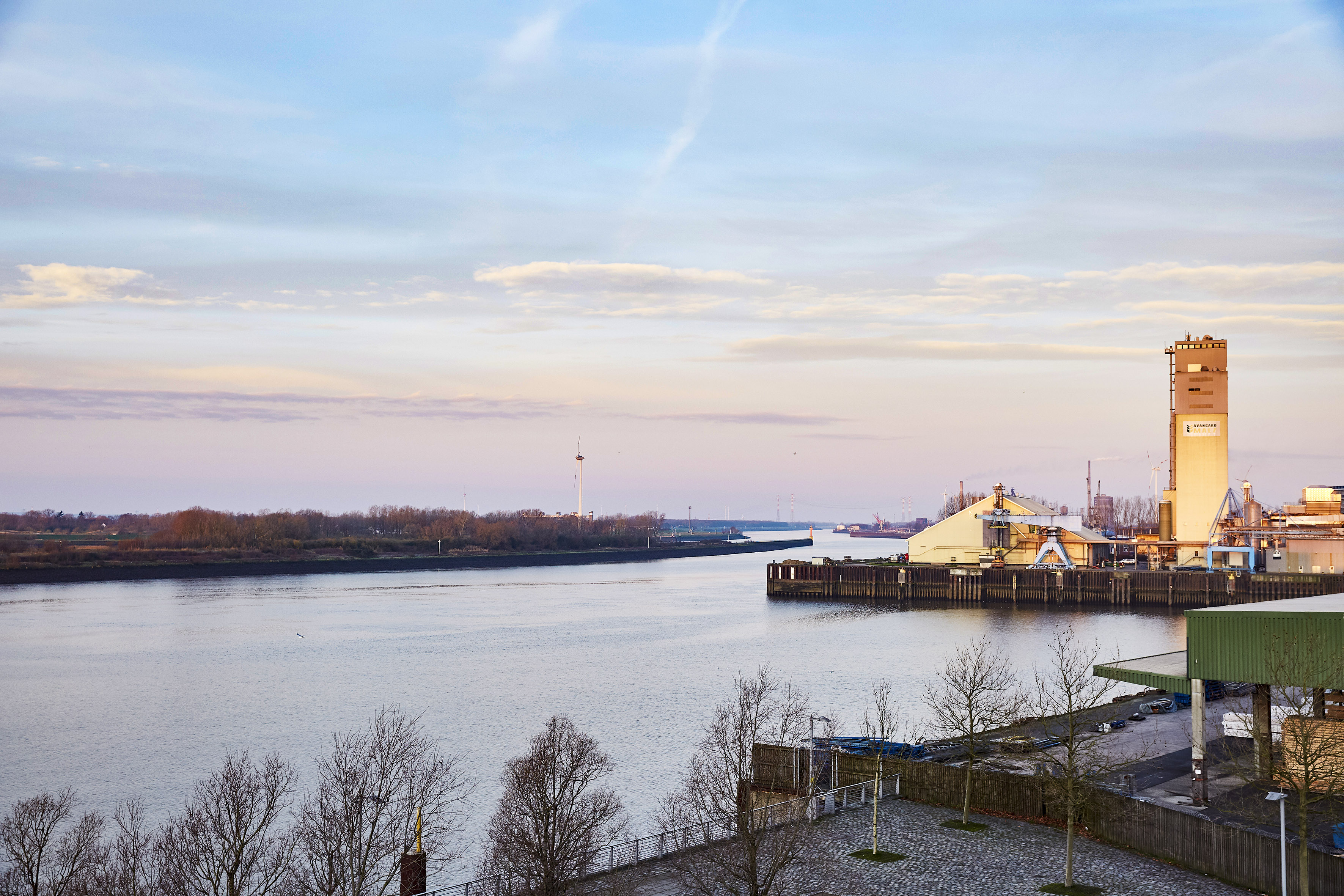 Industrial river port with silos, docks, and winter trees at twilight.
