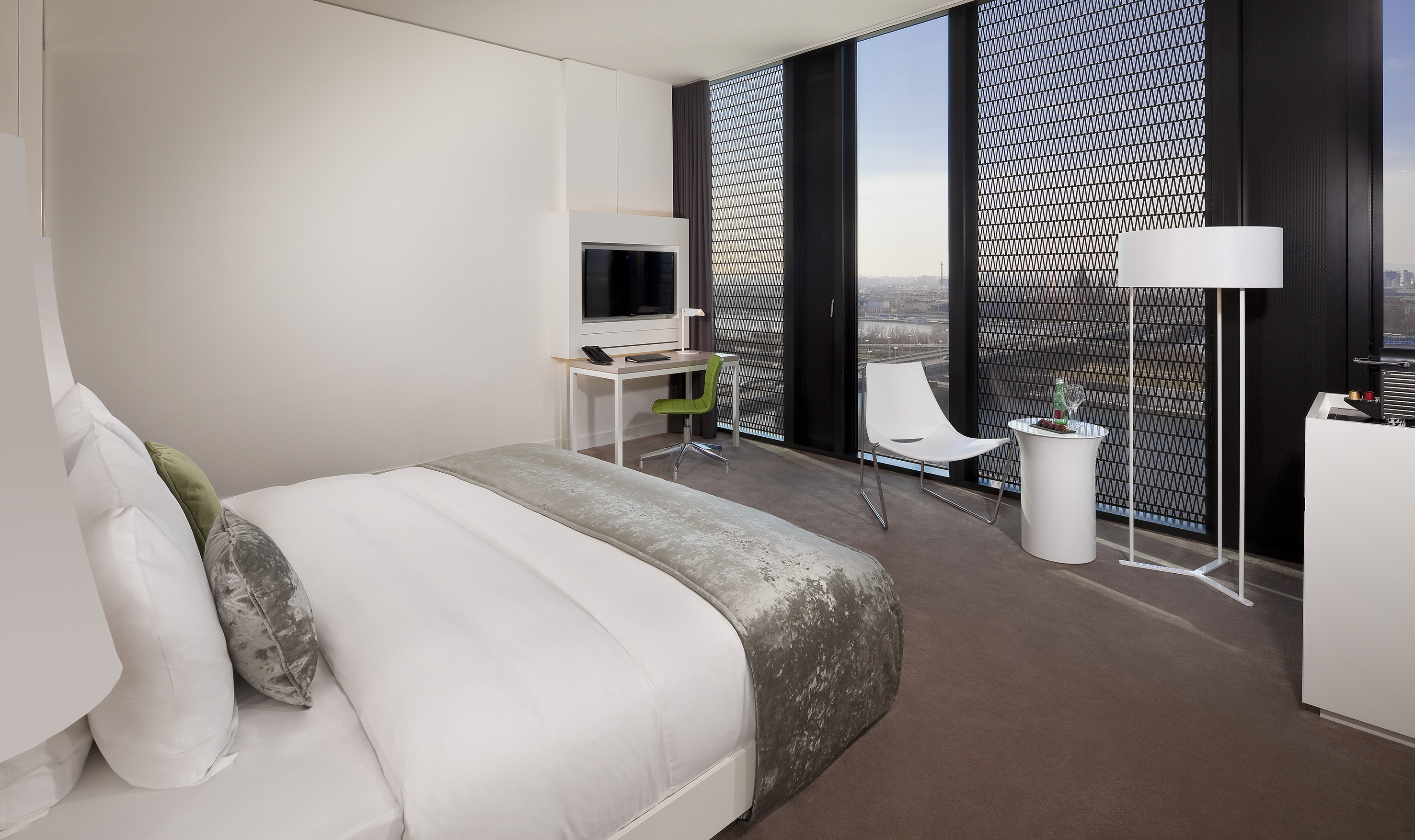 Modern hotel room with bed, desk, and city view from large windows.
