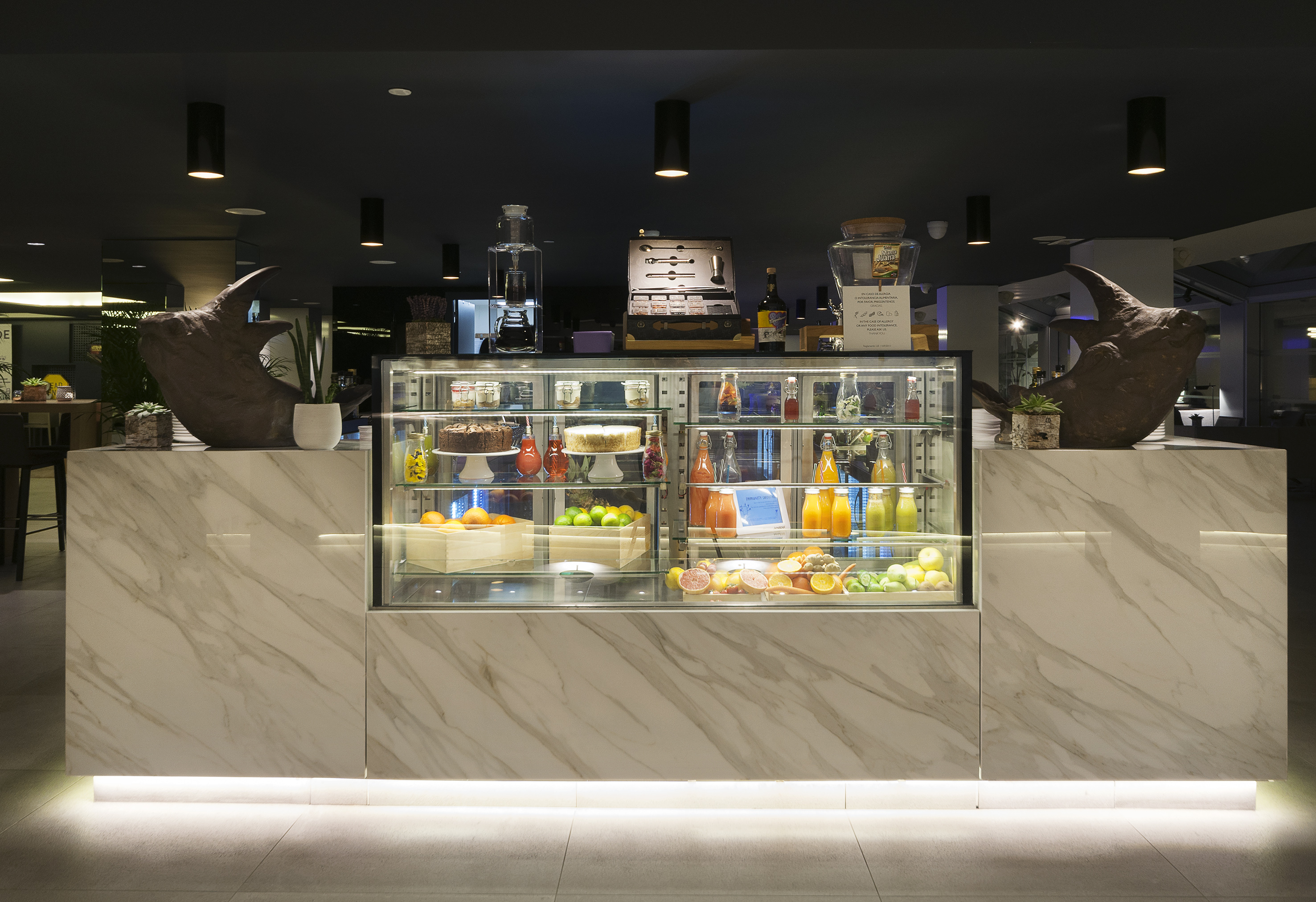 Modern marble counter with fresh juices, cakes, fruit, and decorative sculptures.