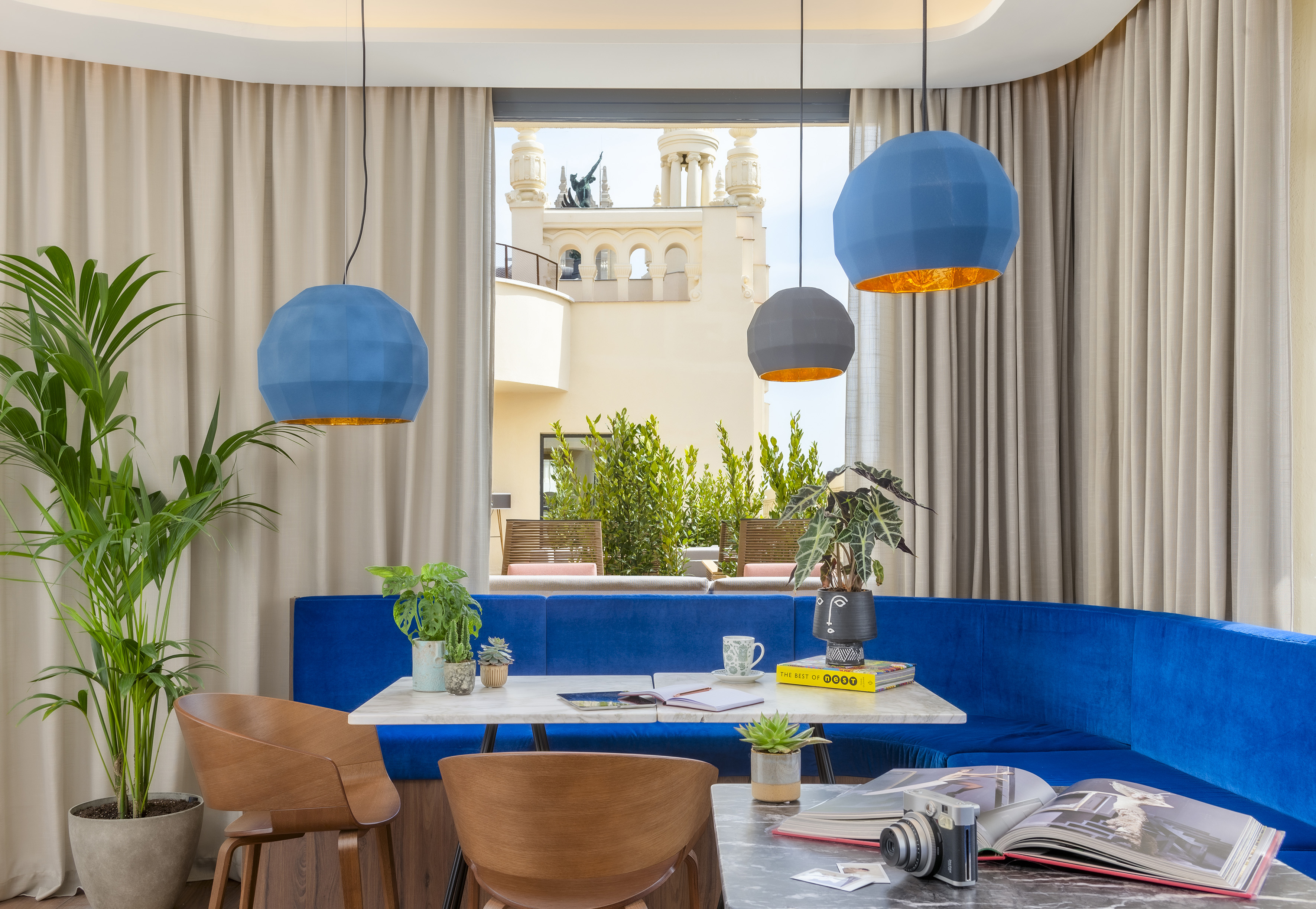 Modern hotel lounge: blue velvet banquette, marble tables, geometric pendant lights.