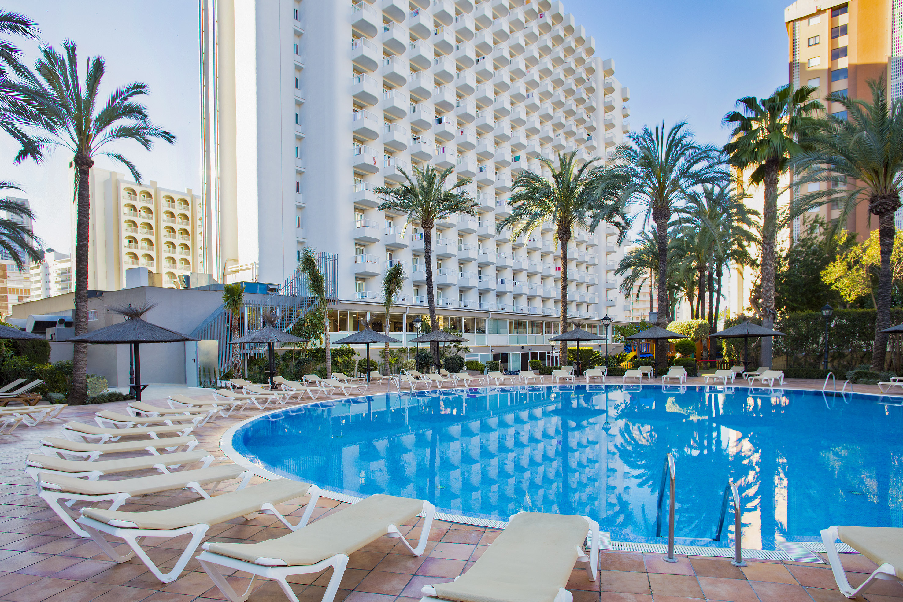 Resort outdoor swimming pool with sun loungers, palm trees, tall hotel.