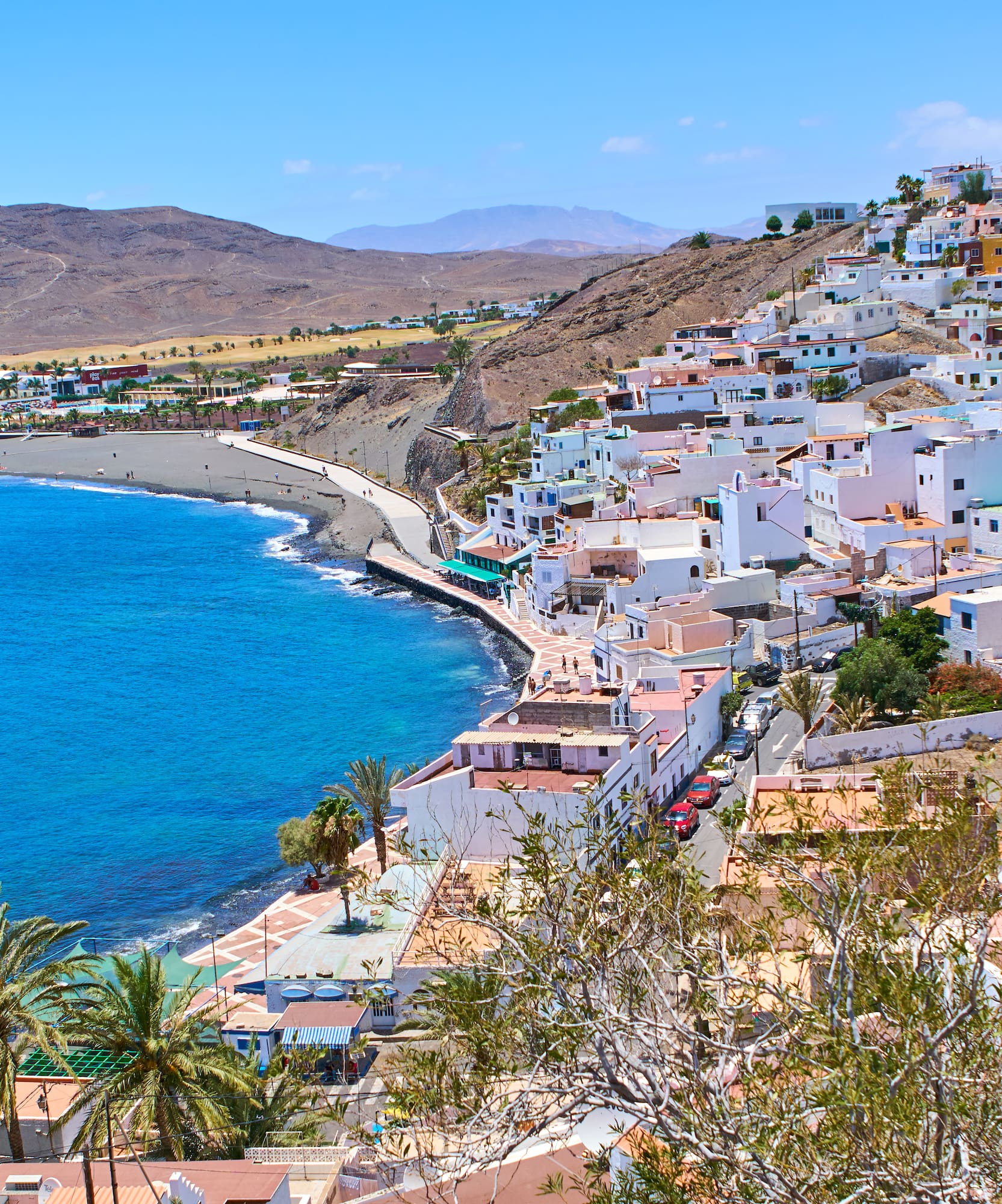 White buildings on a hillside overlooking a bright blue ocean and dark beach.