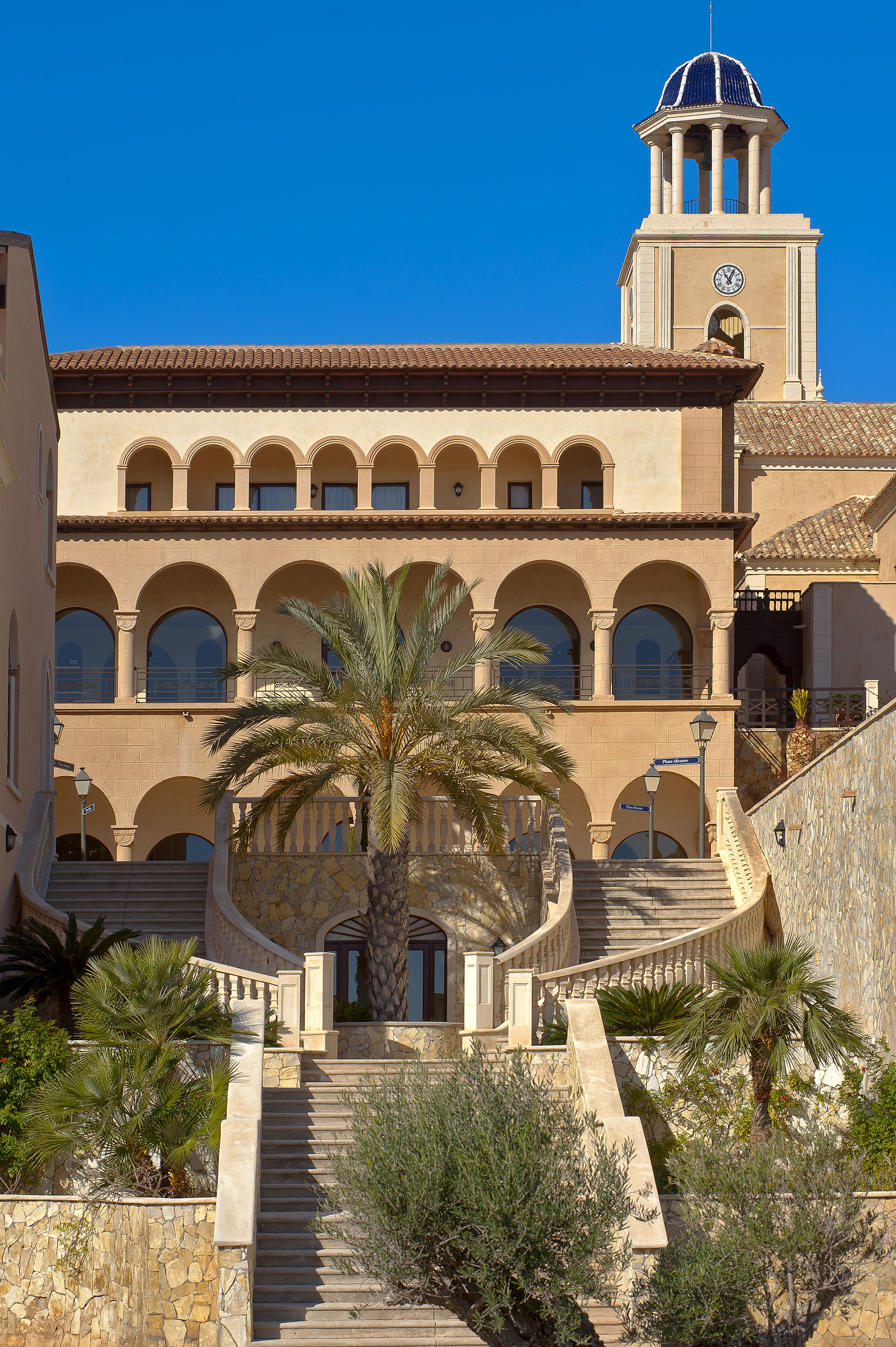 Mediterranean building with arches, clock tower, grand staircase, and palm trees.