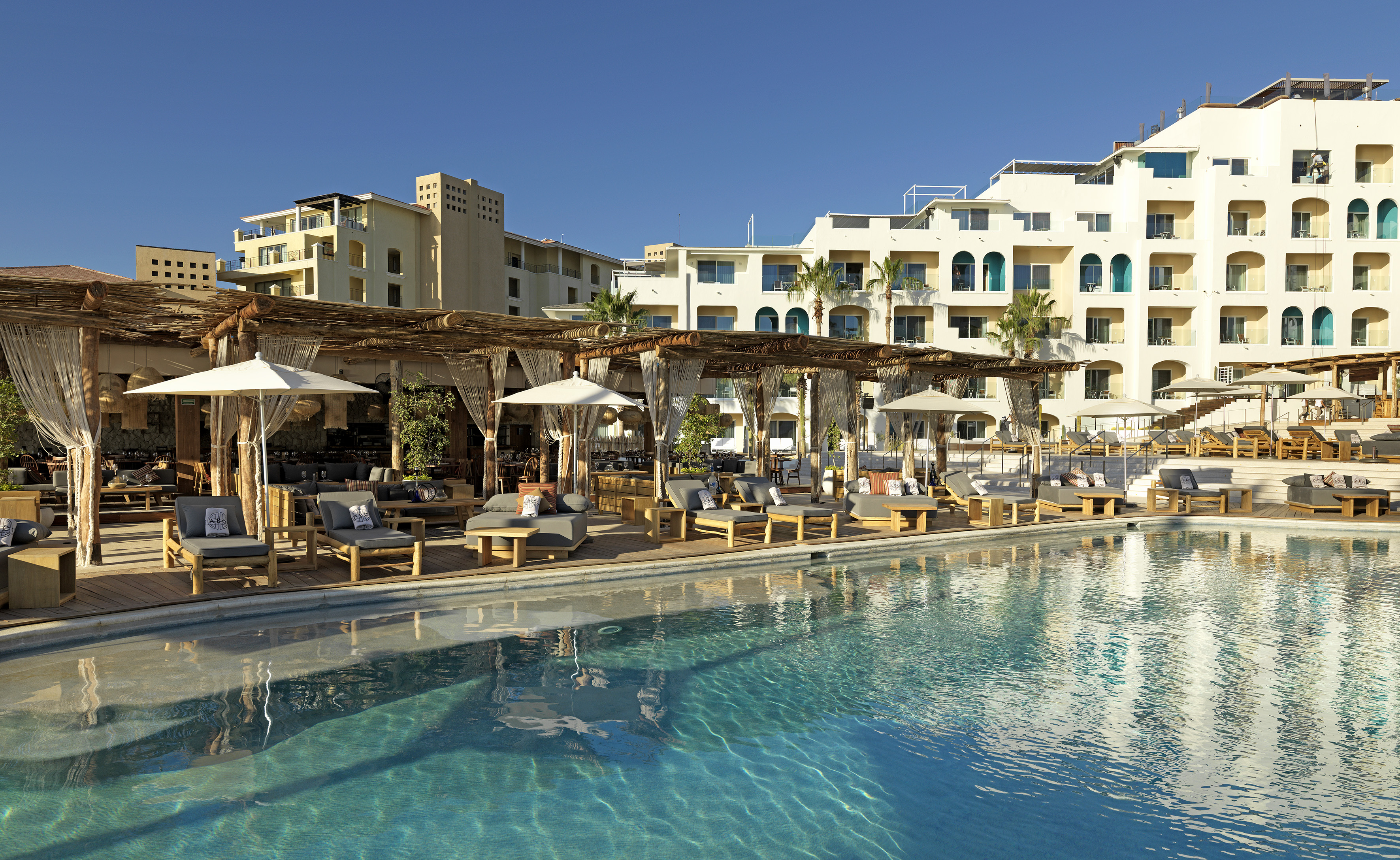Resort pool with modern sunbeds, shaded cabanas, and multi-story hotel buildings.