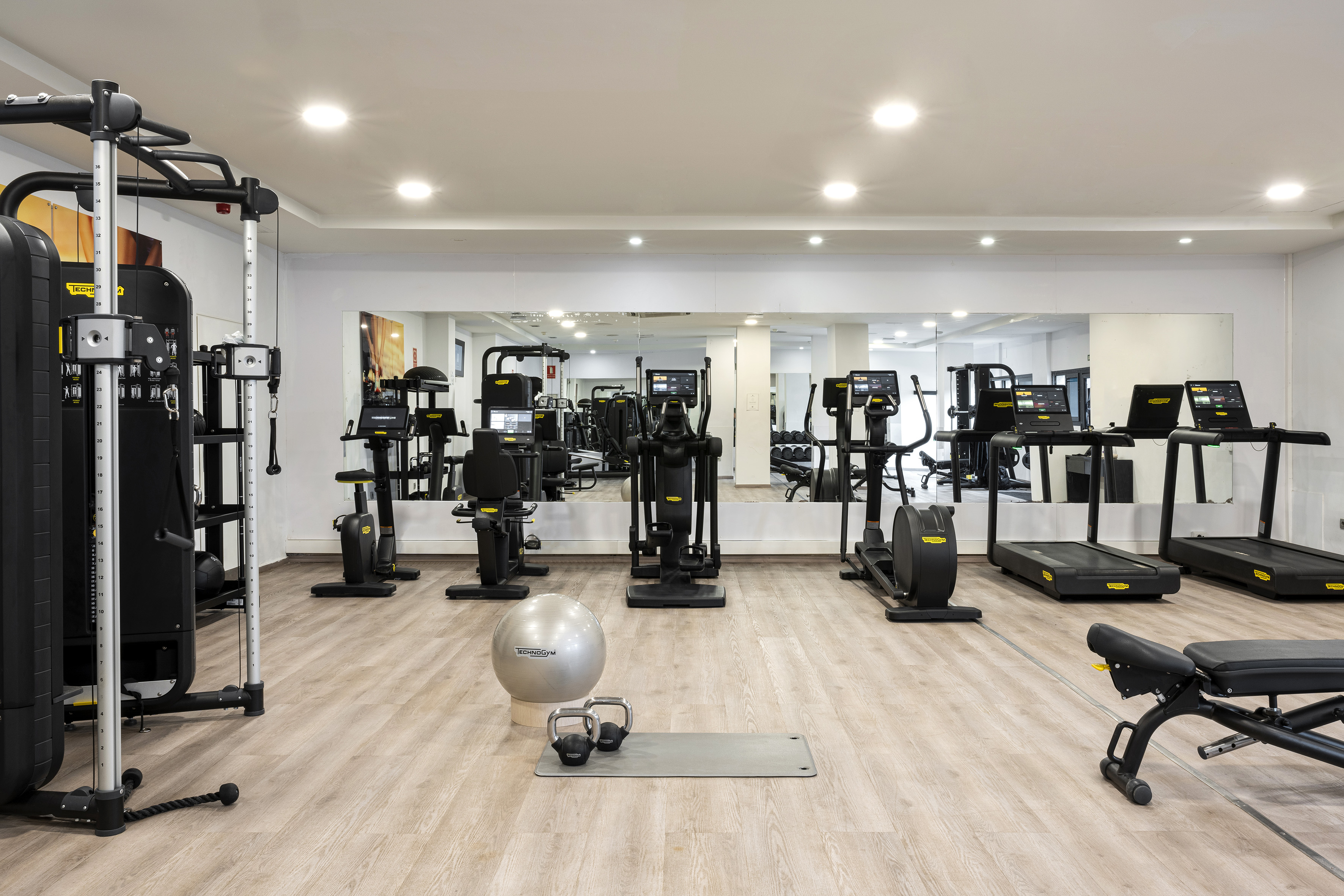 Modern gym with diverse exercise equipment, mirrored walls, and light wooden floor.