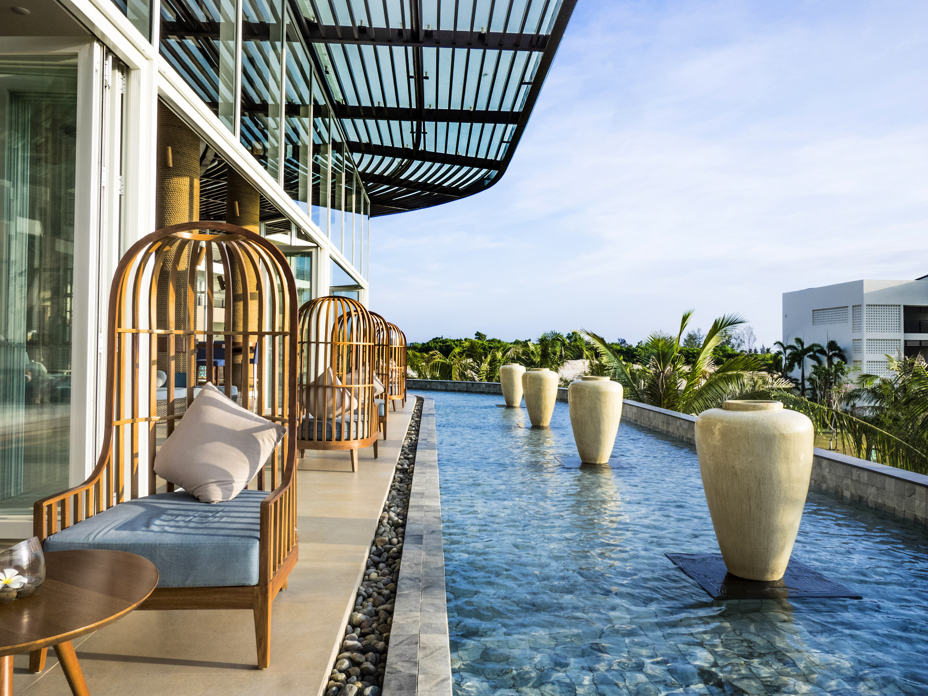 Modern resort patio with wooden lounge chairs, water feature, and tropical palms.