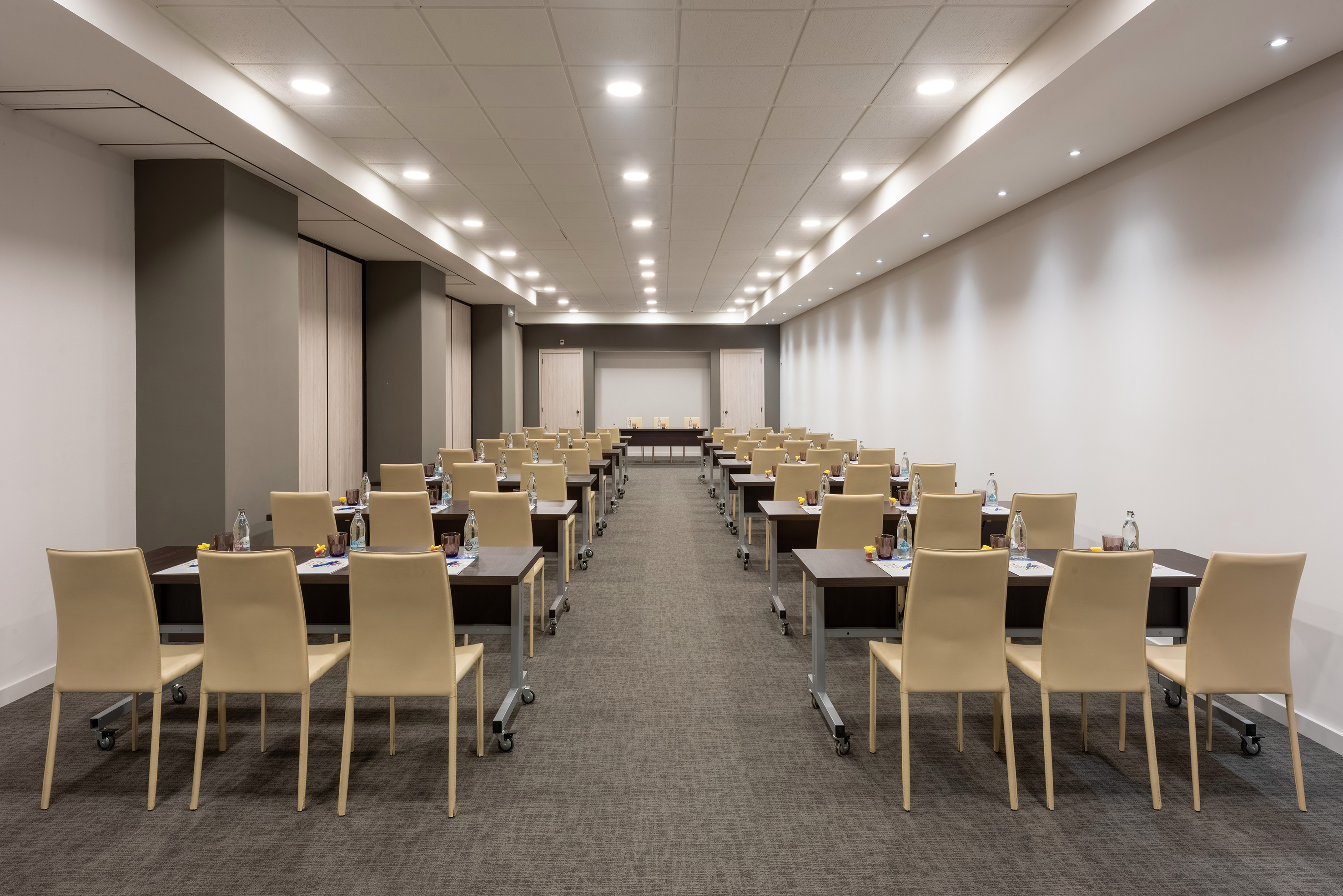 Empty conference room in classroom style with beige chairs, tables, and screen.