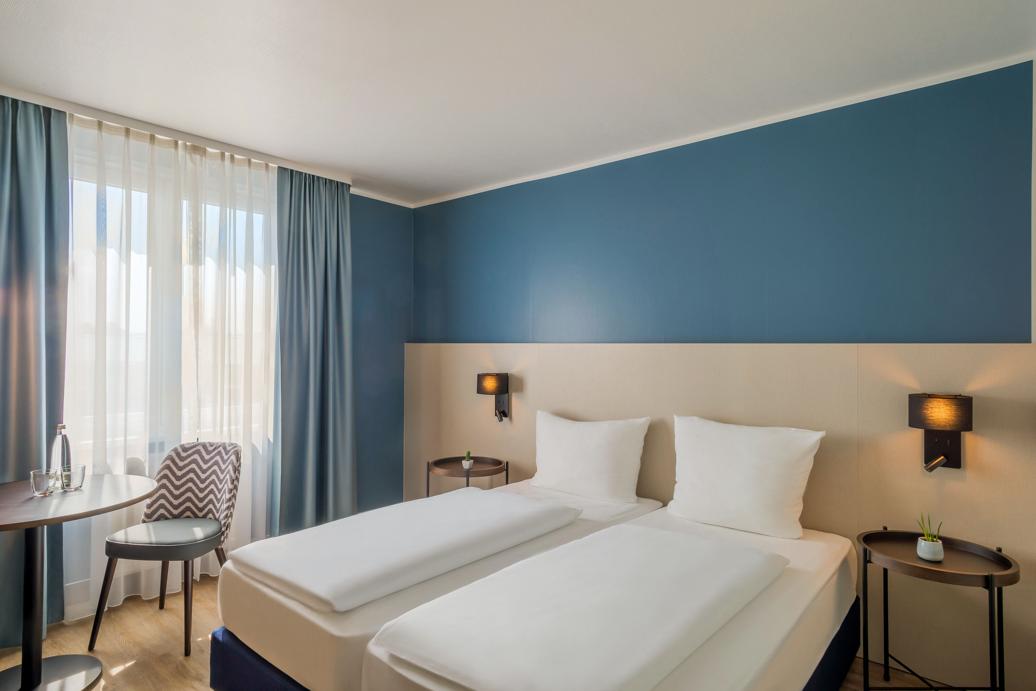 Hotel room with twin beds, blue wall, and window with sheer curtains.