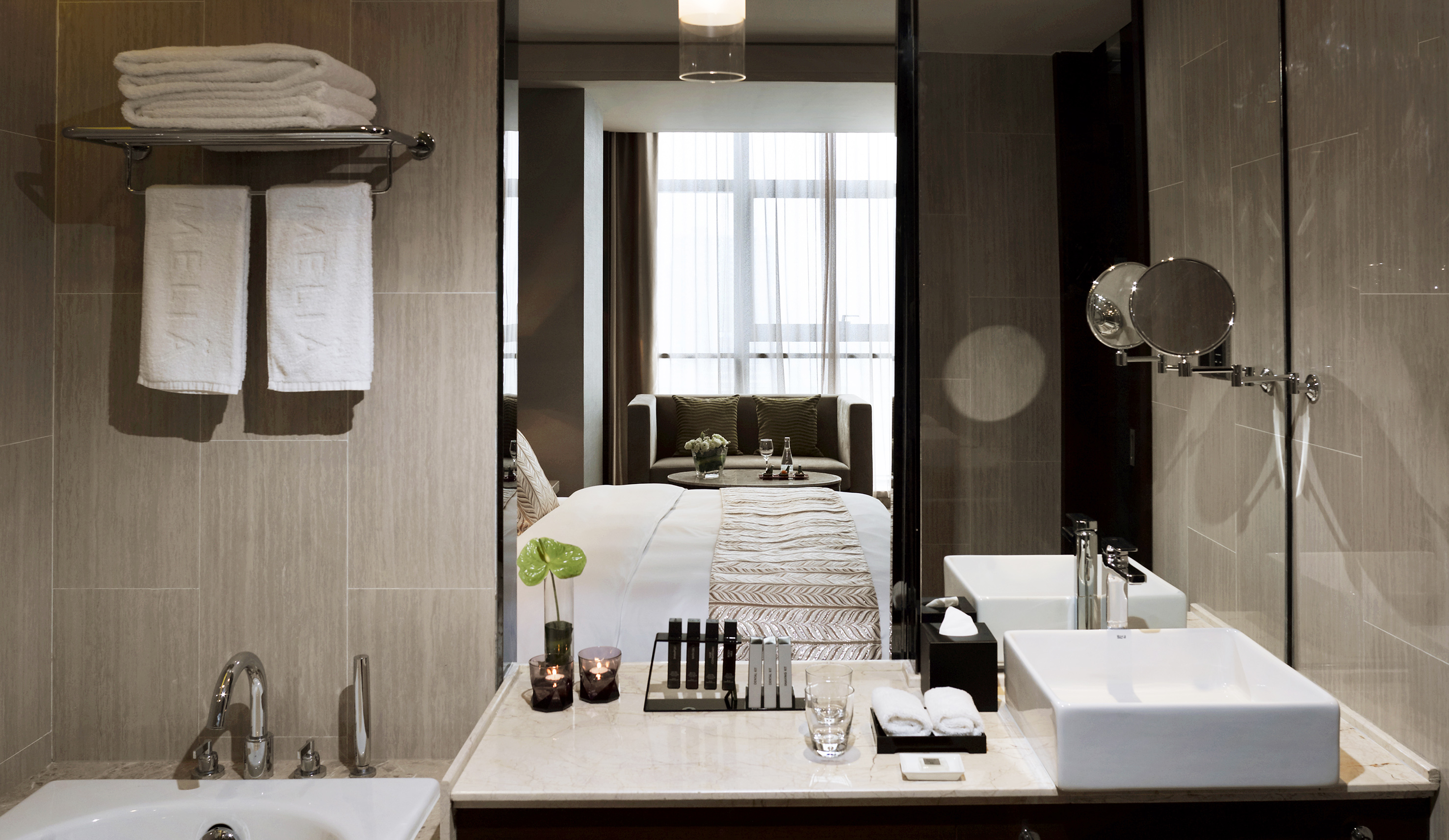 Modern hotel bathroom vanity reflecting bedroom with bed and window.