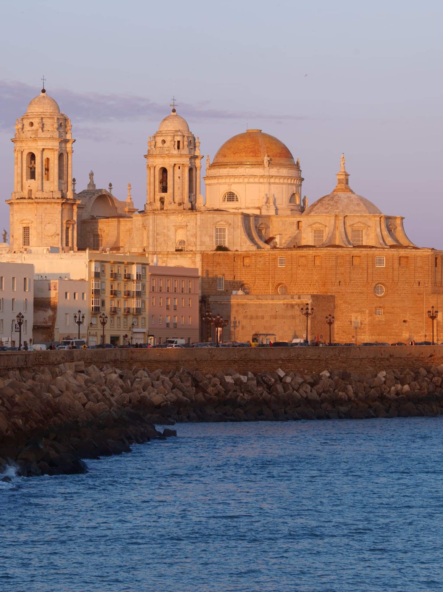 Catedral de Cádiz al atardecer sobre el mar, edificios costeros y rompeolas.