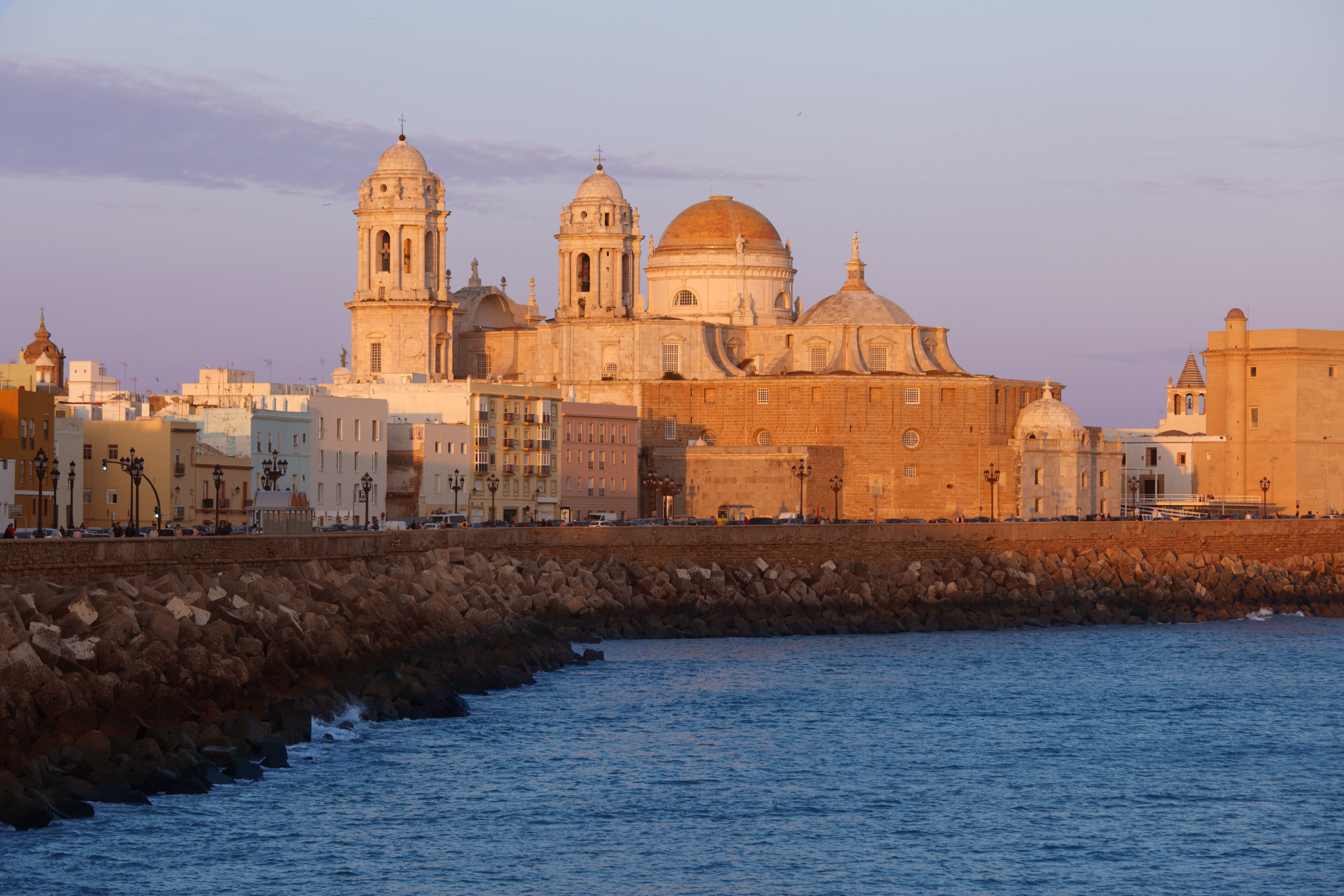 Catedral de Cádiz al atardecer sobre el mar, edificios costeros y rompeolas.
