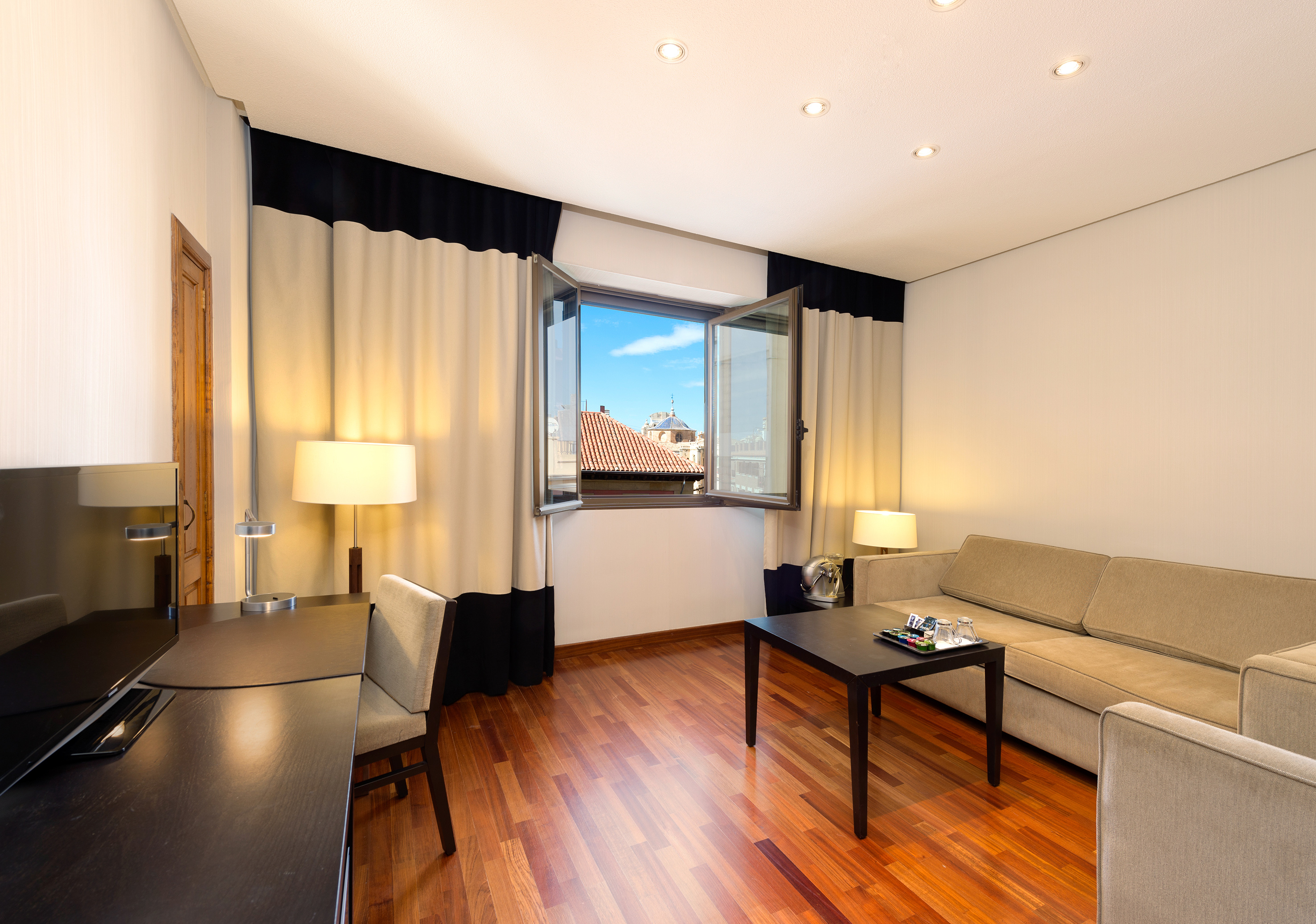Bright hotel suite with sofa, desk, city view from window, and wood floor.