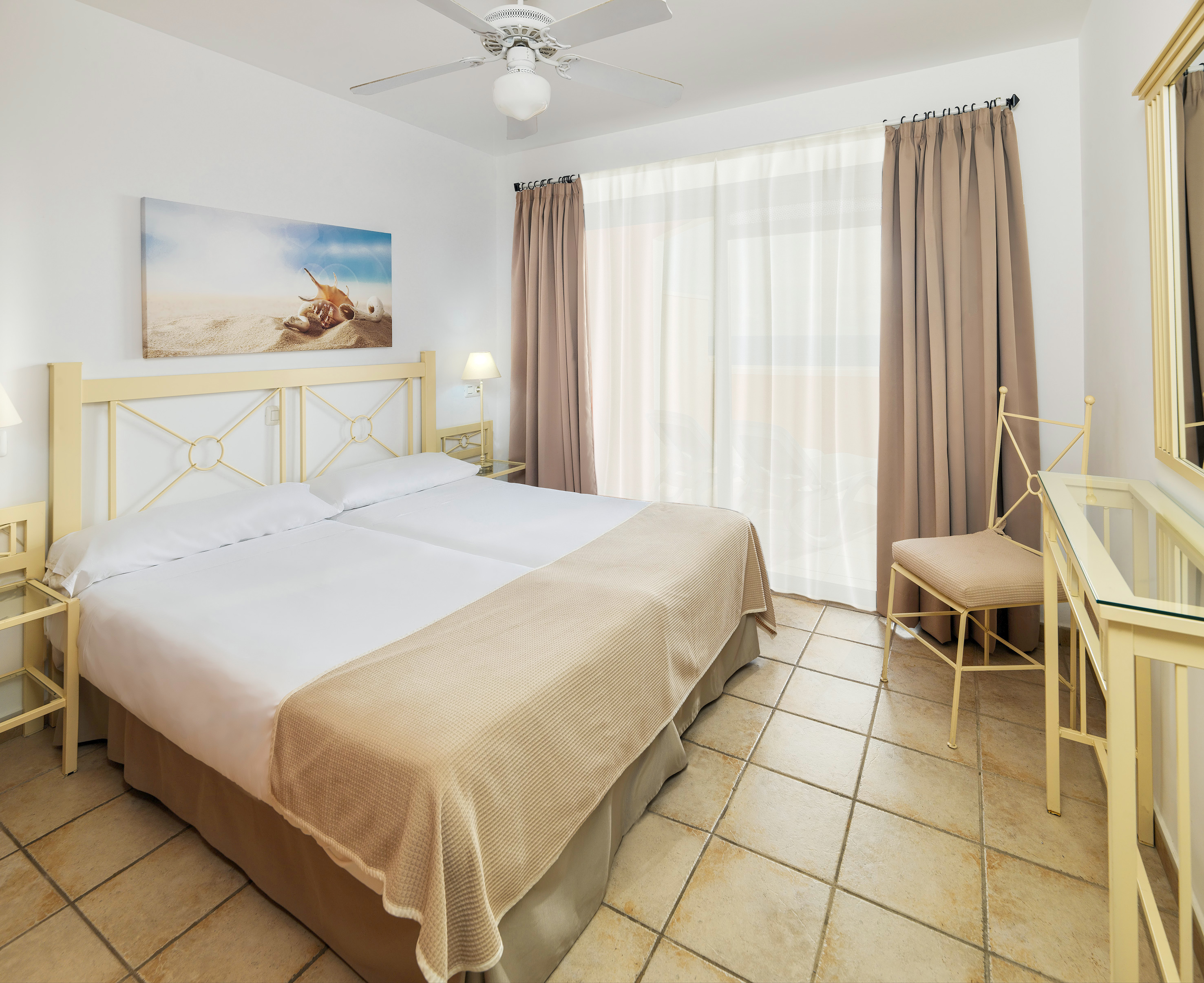 Hotel bedroom with twin beds, beach art, tiled floor, and balcony window.