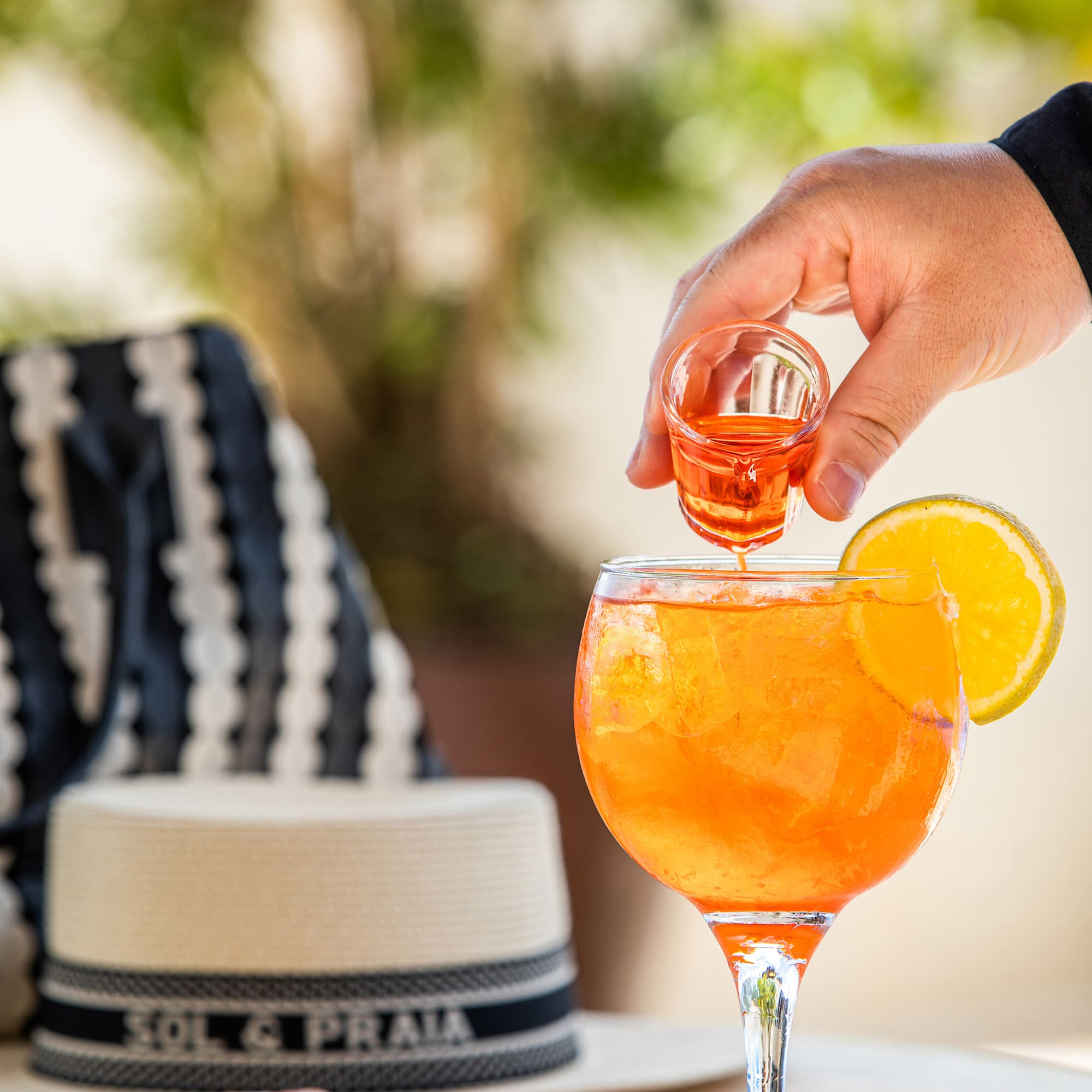 Hand pouring Aperol Spritz cocktail with orange slice; straw hat on table.