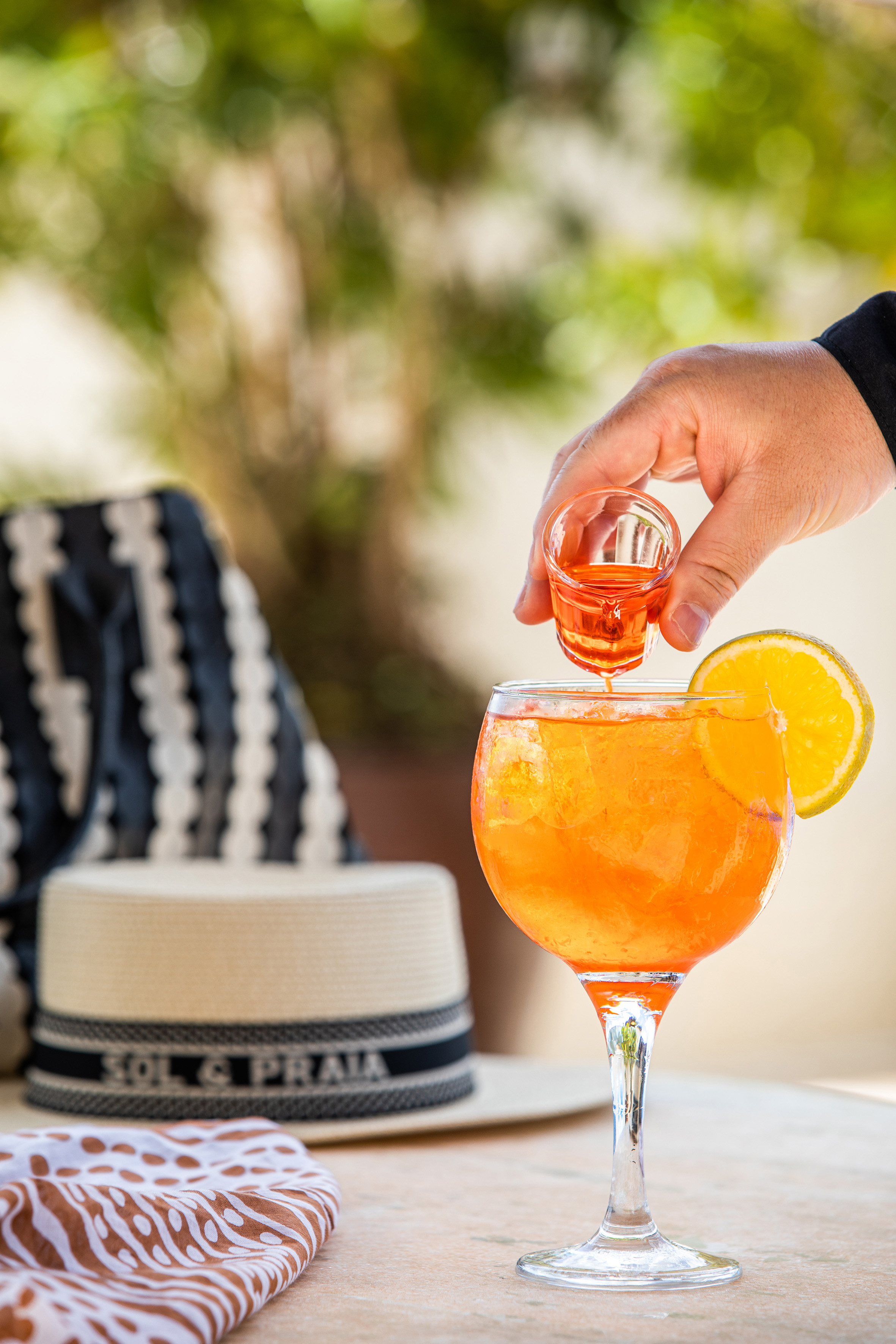Hand pouring Aperol Spritz cocktail with orange slice; straw hat on table.