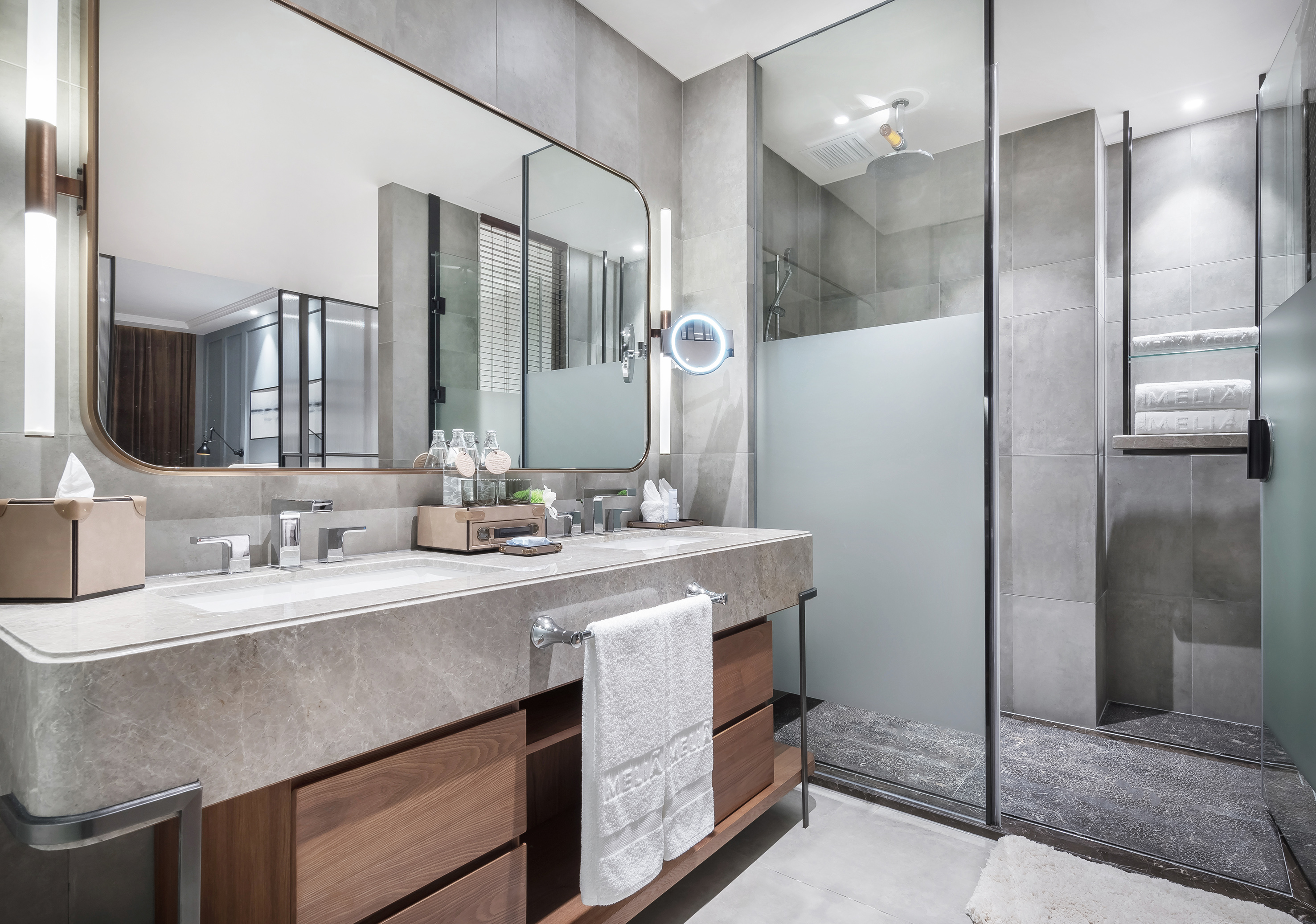 Modern hotel bathroom with double vanity, marble counter, wood cabinets, and glass shower.