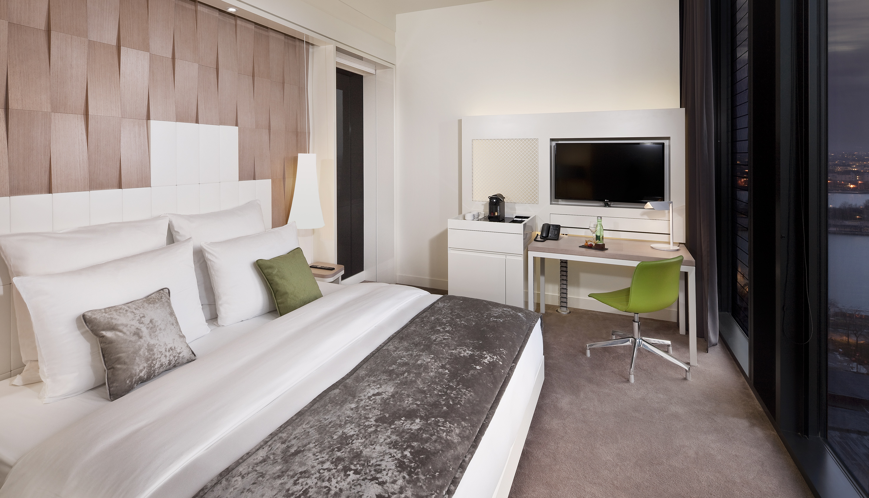 Modern hotel room with bed, workstation, TV, and night city view.