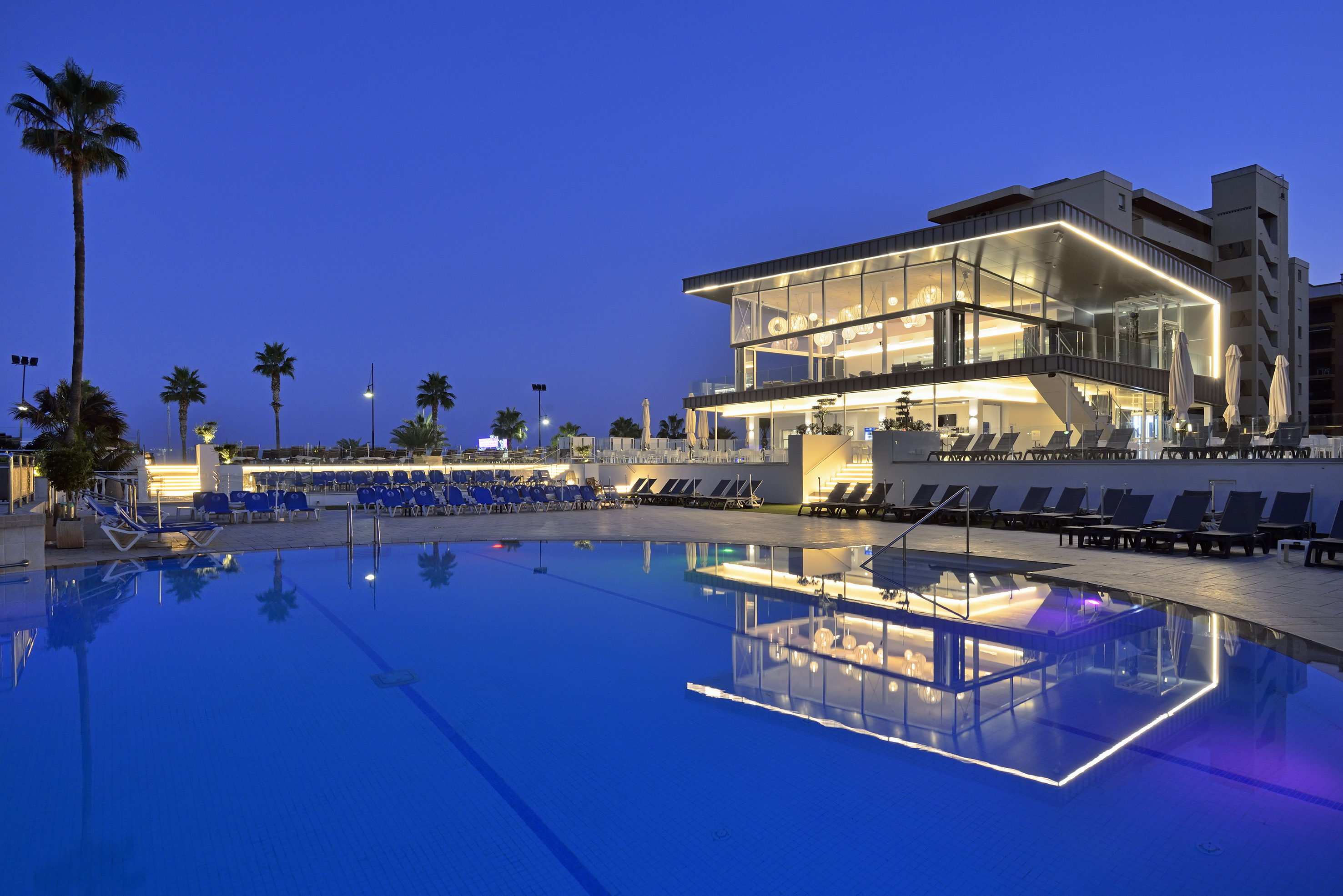 Luxury hotel resort with illuminated swimming pool and palm trees at dusk.