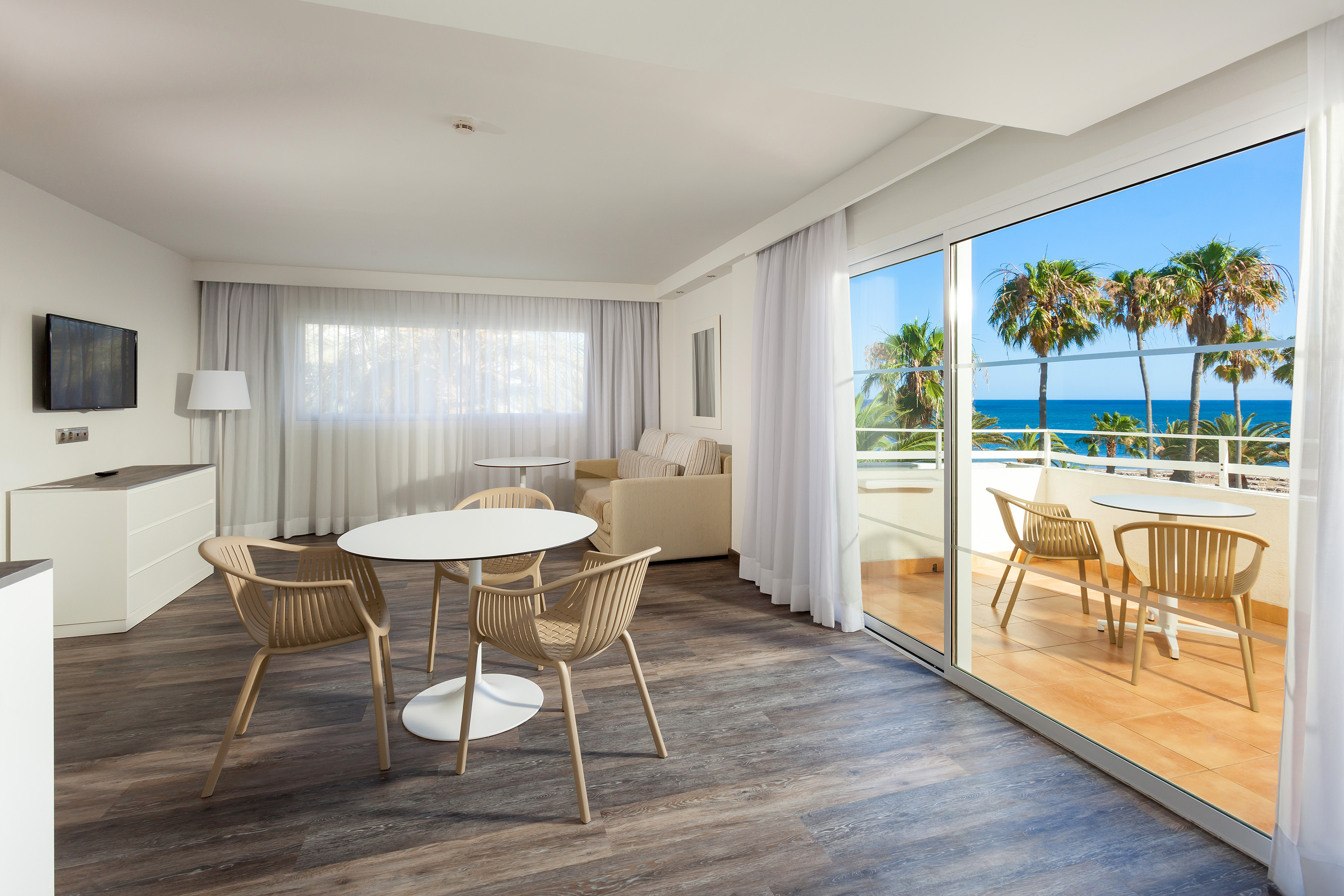 Modern hotel suite living room with ocean view balcony, palm trees, and dining area.