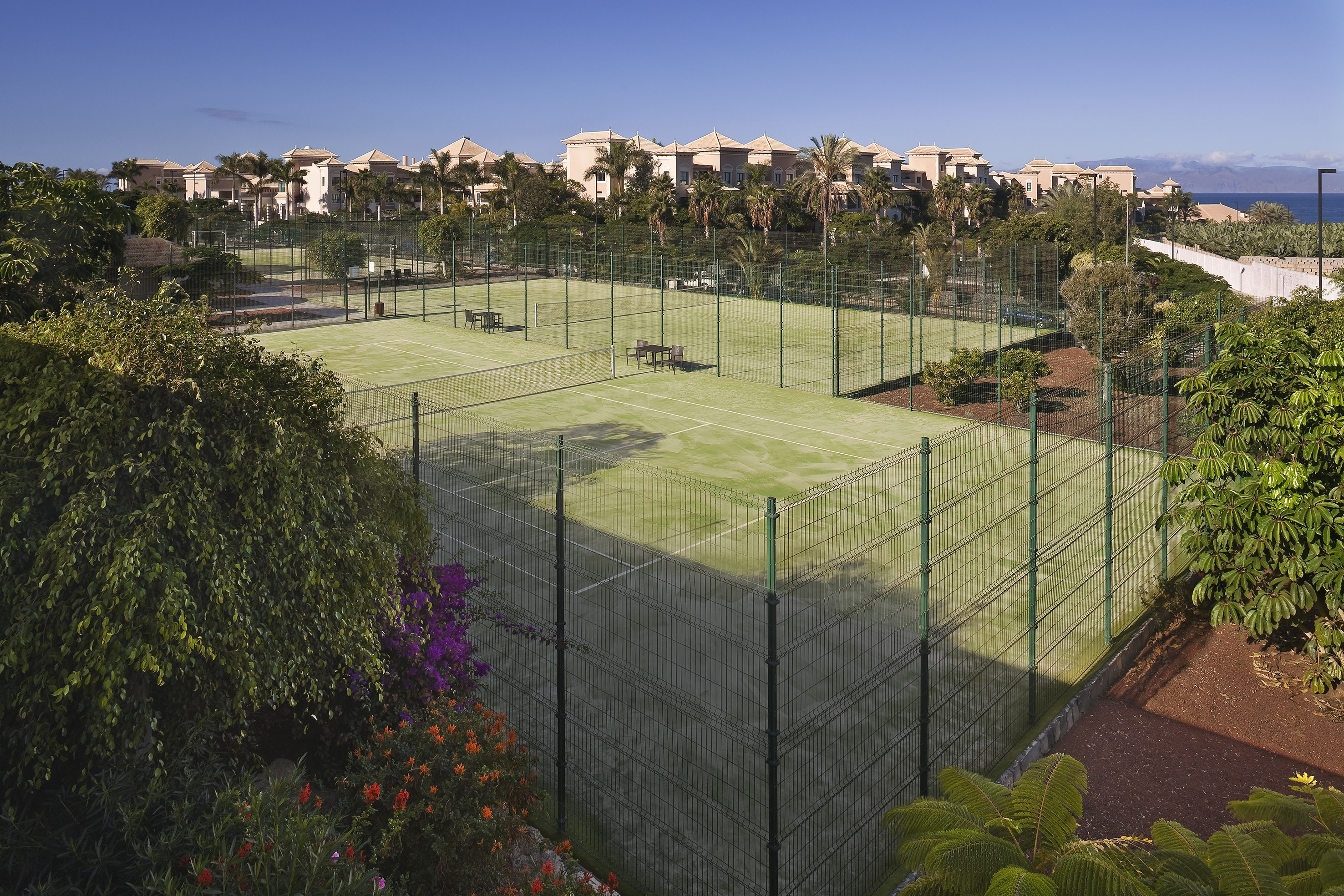 Green outdoor padel courts at a sunny resort with palm trees and buildings.
