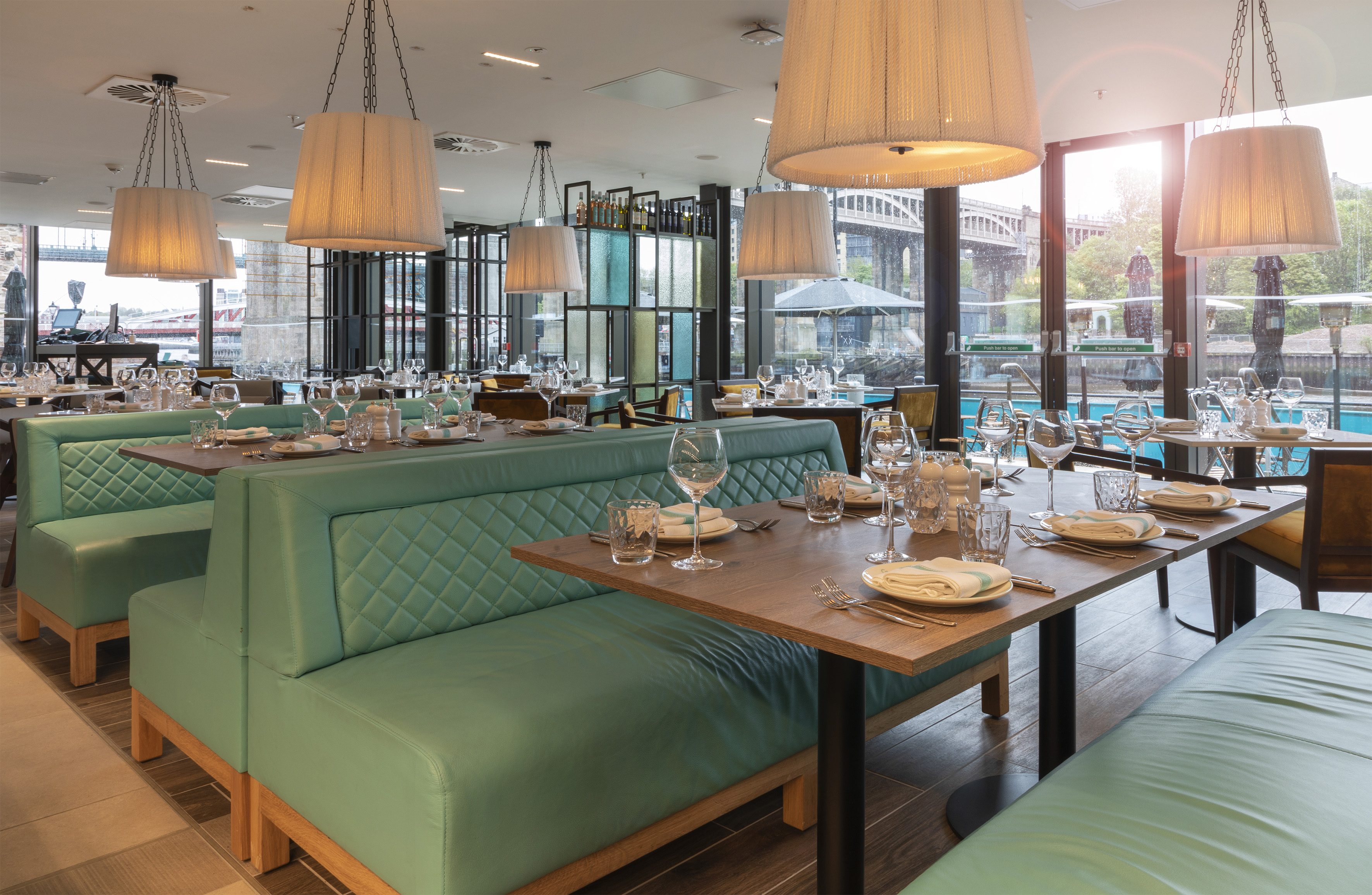 Modern restaurant interior with teal booths, set tables, and a sunny window view.